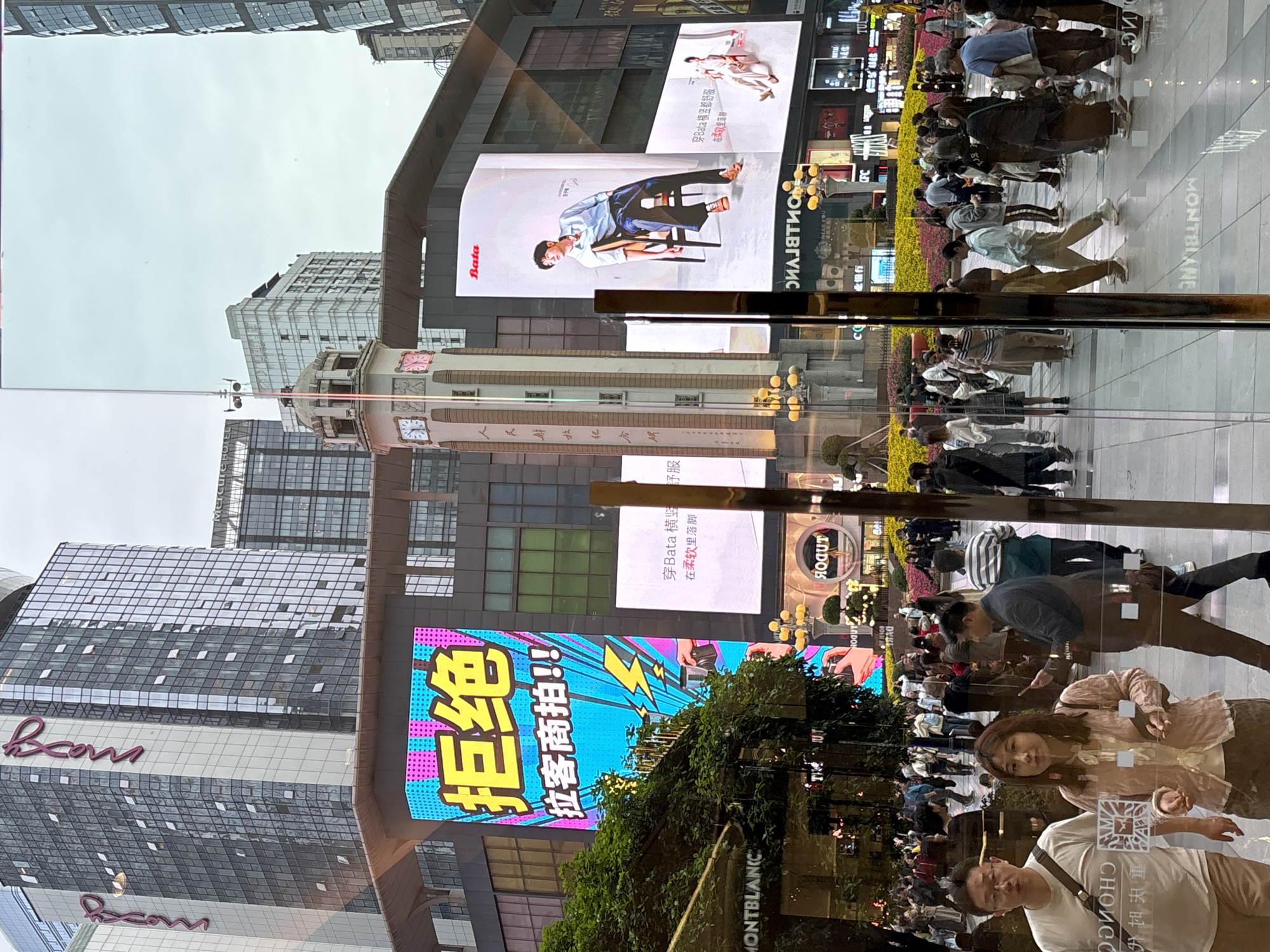 The image shows a street scene in Yuzhong District, China, reflecting off of glass or a window. The reflection includes people walking on the street, along with various storefronts and advertisements. There's a crowd of people, including a woman looking at the camera.
Visible store signs include "MONTBLANC," "Boom," and "Bata" (with advertisements featuring models). There is also a sign in Chinese, translated as "Reject unauthorized commercial photography!" Other signs are partially visible, showing Chinese characters and English letters.
The buildings reflected in the glass are modern high-rises, with a mix of glass and solid facades. One older building with a clock tower is also visible. The sky is overcast, indicating cloudy weather.