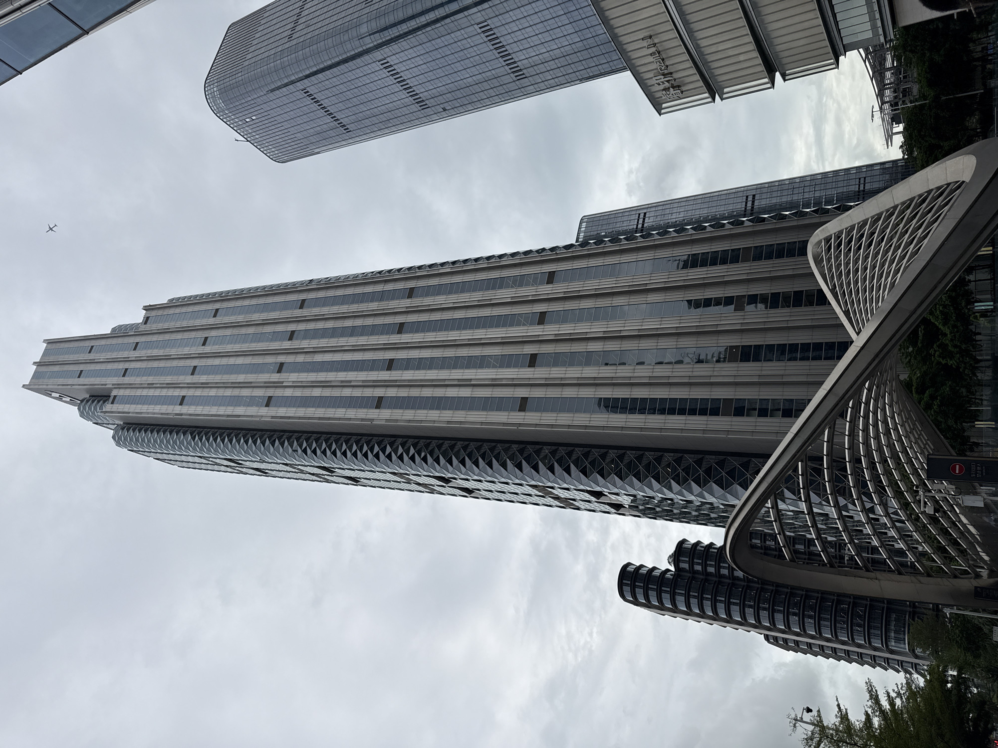 The image shows a cluster of modern skyscrapers in Nanshan District, China, under a cloudy sky. The central building is a tall, narrow skyscraper with a distinctive facade featuring vertical metallic elements and diagonal diamond-shaped patterns. To its left, a shorter building with a curved, segmented design is visible, and to its right, another taller skyscraper with a grid-like glass exterior. In the foreground, a futuristic, arching metal structure spans across the lower part of the frame, creating a canopy-like effect. An airplane is visible in the sky above the buildings. The environment appears to be an urban setting with some trees on the left side of the image. There are no people clearly discernible in the scene.