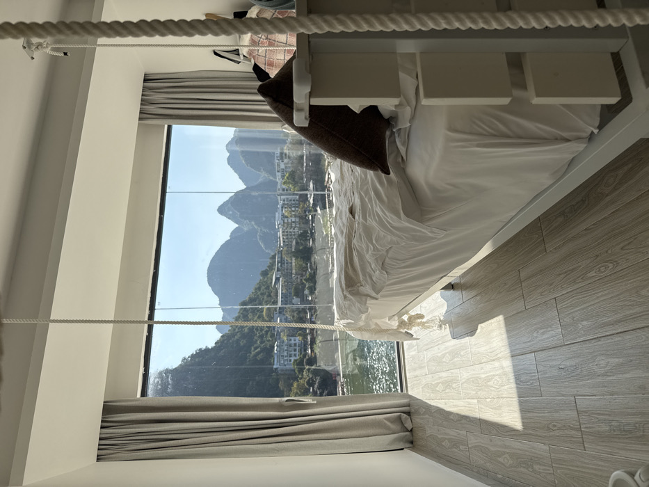 The image displays a minimalist bedroom interior in Guilin, China, featuring a panoramic, floor-to-ceiling glass window that overlooks a river, urban buildings, and characteristic karst mountains under a clear, bright sky. The room is illuminated by direct sunlight, which reflects off the water and casts shadows on the light wood-effect tiled floor. A white, unmade bed with crumpled white linens and a single dark brown throw pillow occupies the center-right foreground. The bed frame is white, featuring vertical slat detailing on the visible side. Thick, braided white ropes descend from the white ceiling, with one visible on the left side of the bed, terminating in a decorative knot, and another partially visible on the far right, secured by a metal ceiling hook. Light grey curtains are partially drawn on both sides of the expansive window. No individuals are present in the scene.