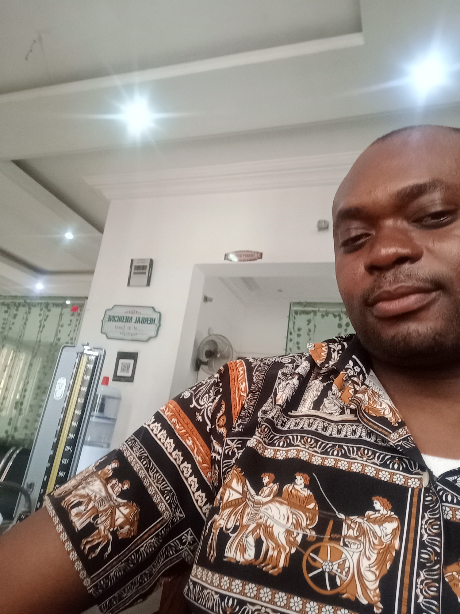 A man is captured in a selfie or close-up shot within an indoor setting, likely a clinic or healthcare facility in Gwarinpa, Nigeria. He is a dark-skinned individual, appearing to be in his 30s or 40s, with a shaved head or very short hair and stubble on his face. He looks directly at the camera with a neutral expression, possibly a slight smirk. He wears a short-sleeved, collared shirt featuring a distinctive black, brown, and white pattern depicting classical or ancient figures, some riding horses and chariots.
The background reveals a well-lit room with white walls and a white ceiling adorned with recessed lights and decorative crown molding. One bright light source causes a prominent lens flare in the upper left. To the left of the man, medical equipment is visible, including a tall, grey manual blood pressure monitor with a vertical scale showing numbers like 0, 100, 150, 200, 250, and 300. A water dispenser with a red top and clear bottle is partially seen nearby.
On the wall behind the man, a sign is prominently displayed, which appears reversed, indicating it might be a reflection or seen through a transparent medium. The legible, reversed text reads "HERBAL MEDICINE" and below it, partially visible, "lead us to...". A small framed item and a square QR code poster are also on the wall below the main sign. Higher up, a digital clock or display unit is visible. On the right wall, a small, wall-mounted oscillating fan is partially obscured. Green patterned curtains are visible through a doorway or window on the far left of the frame. The overall impression is a clean and functional interior, likely a professional waiting area or consultation room.