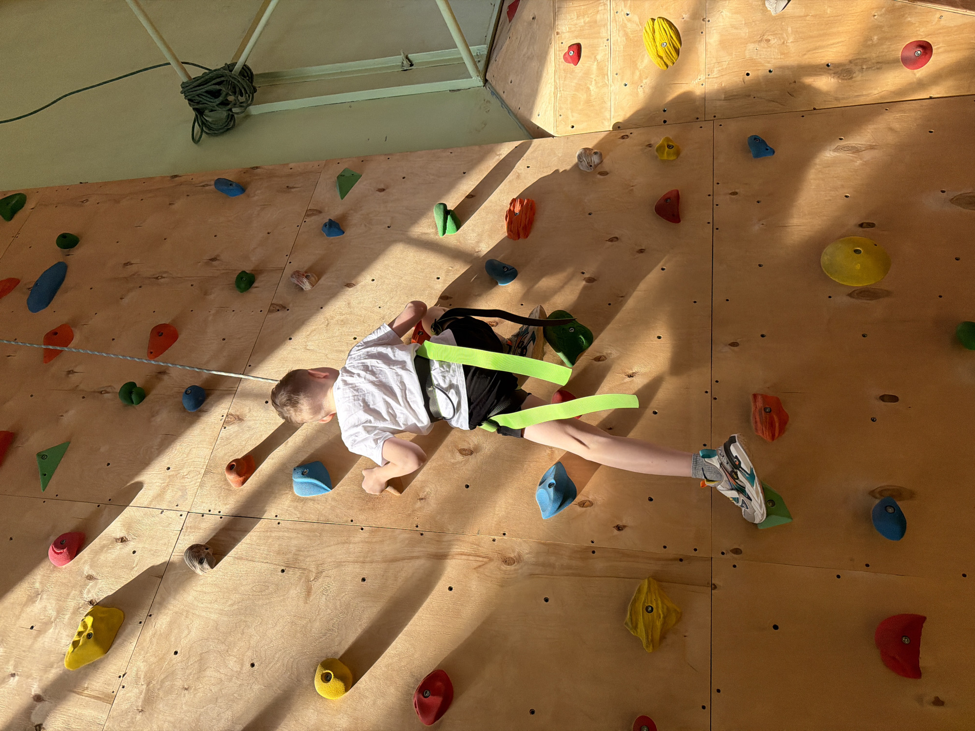 A young boy, secured with a climbing harness and lime green safety straps, lies horizontally against a climbing wall. He wears a white shirt, black shorts, and athletic shoes with gray and turquoise accents. The wall is constructed of plywood and features various colored climbing holds (red, yellow, green, orange, blue, and gray) scattered across its surface. Light streams in from the upper right, casting shadows across the wall. A coiled rope hangs from the ceiling in the upper left corner. The location is Izhevsk, Russia.