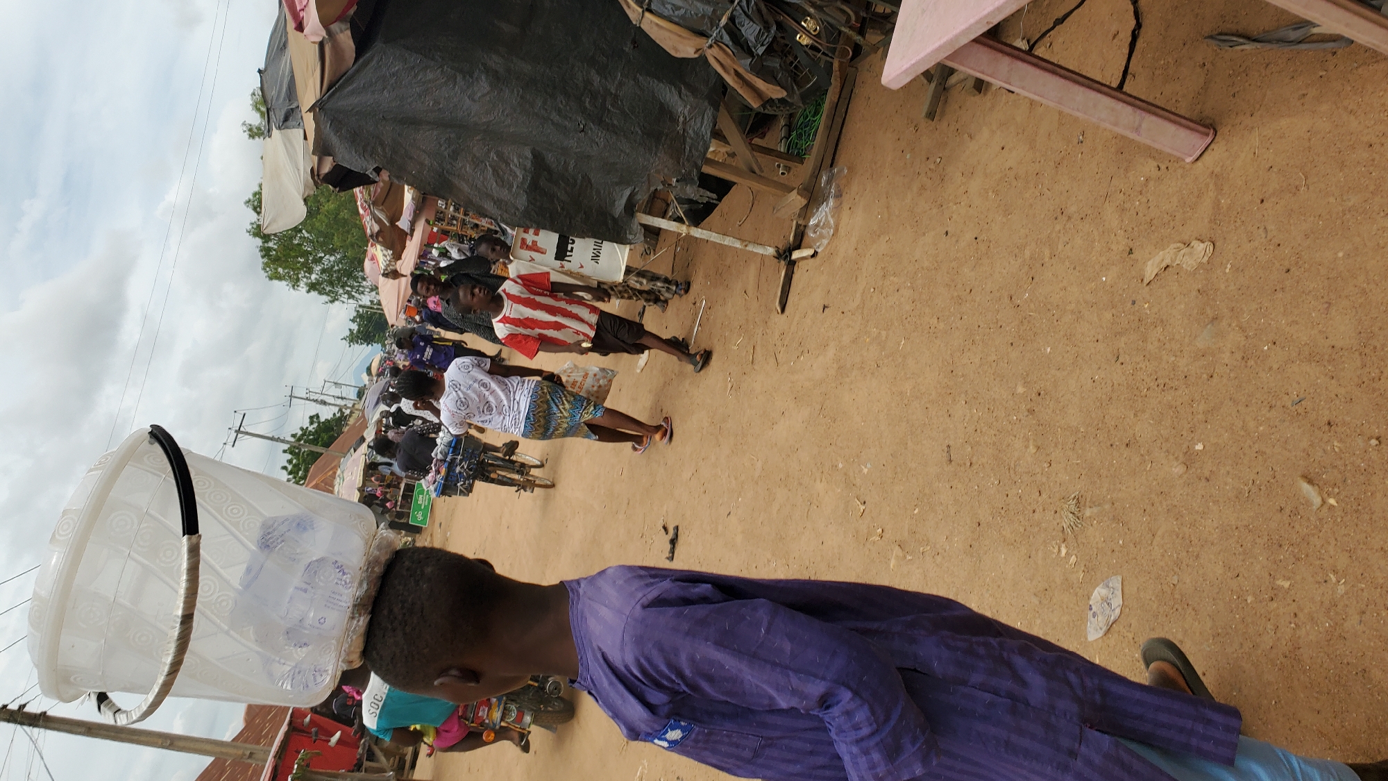 The image captures a bustling street scene in Jalingo, Nigeria, likely a market area, on an overcast day. In the foreground, a young boy wearing a traditional purple tunic and light blue trousers stands facing away from the camera, balancing a large, clear plastic bucket filled with what appear to be water bottles on his head. The bucket has a black handle wrapped in white tape.
The background reveals a busy street with many people walking and interacting. To the left, a person on a motorcycle is visible. Several market stalls with colorful awnings are set up along the street, with various goods displayed. People are seen walking, some carrying bags. A woman in a white patterned top and a patterned skirt is walking away from the camera, holding a plastic bag. Further in the background, more individuals and stalls are present, with trees and buildings lining the street. The ground is unpaved, appearing to be dirt or sand, and scattered with some debris. A pink plastic table is visible in the lower right corner of the image. The overall atmosphere suggests a lively commercial and social hub.