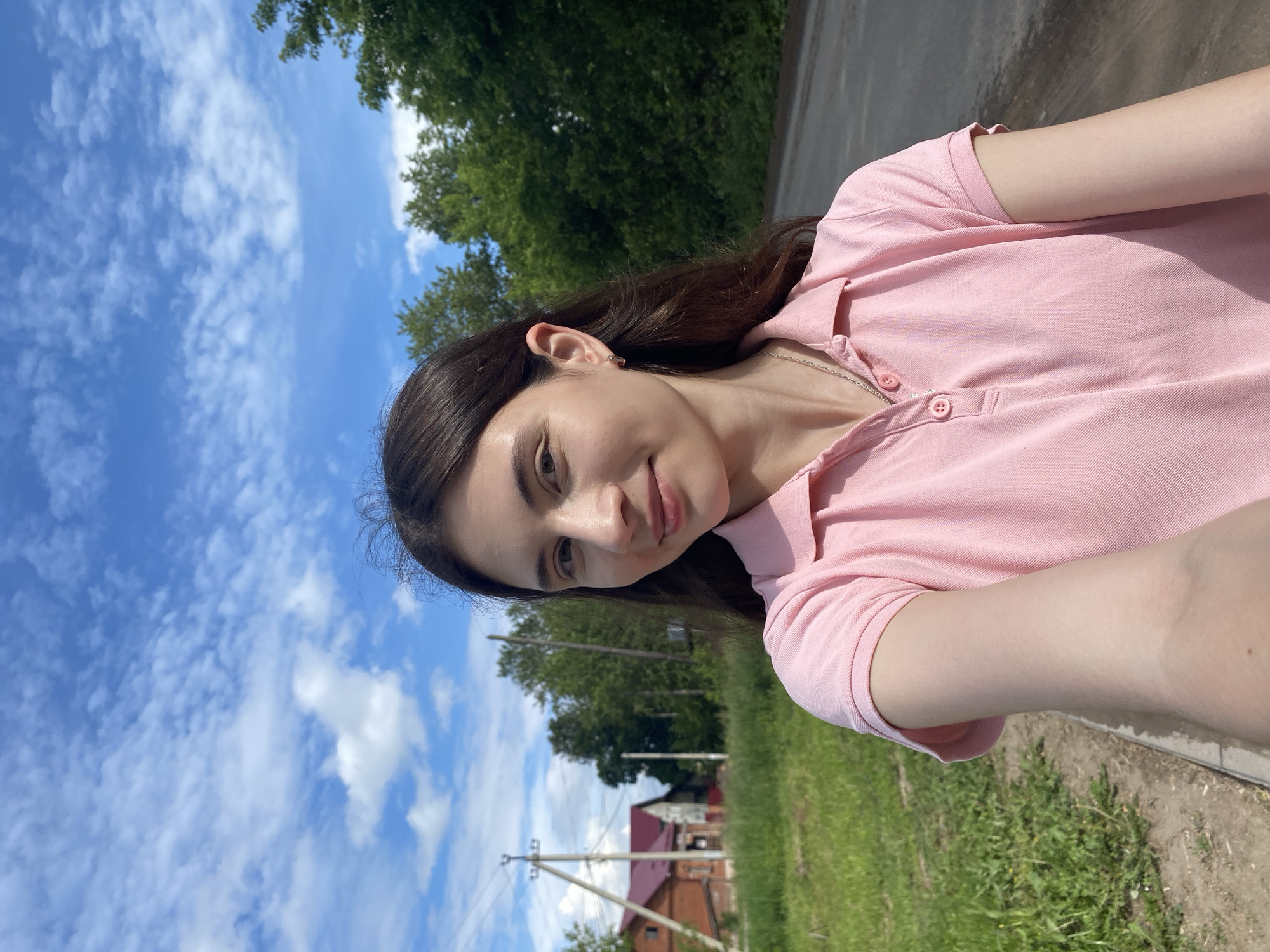 A young woman with long, dark hair is taking a selfie outdoors. She is wearing a pink polo shirt and a silver necklace. She is standing in front of a rural landscape with a road, grassy fields, trees, and a few houses with red roofs. The sky is blue with scattered white clouds. The scene appears to be in the city of Votkinsk, Russia, based on the typical landscape and architecture found in the region.