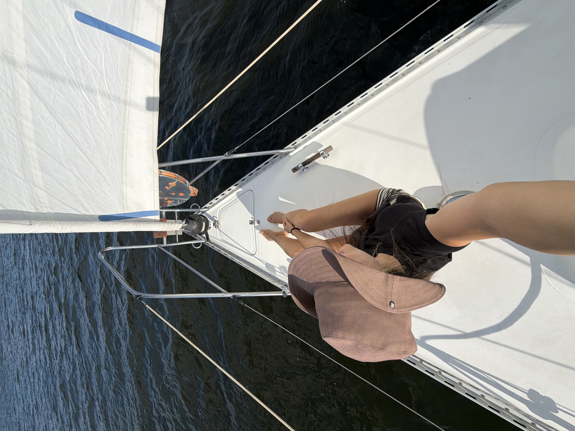 The image shows a person, likely female, sitting on the bow of a white sailboat. She is wearing a wide-brimmed brown hat, a black t-shirt, and striped shorts. Her legs are extended forward, and her bare feet are visible. A white sail is visible in the upper right portion of the image, and the dark blue water of a lake or river surrounds the boat. The image appears to be taken from a high angle, looking down at the person and the boat. There is no indication of the city of Kamyshin, Russia, or any other specific location.