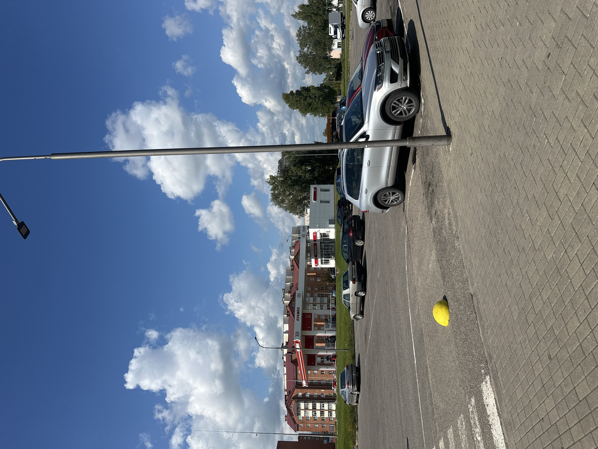 The image depicts a gas station with the Lukoil branding visible, situated in the city of Maladzyechna, Belarus. In the foreground, a silver SUV is parked in a paved lot, with a red vehicle partially visible next to it. Other cars are also parked in the lot. A tall lamp post stands prominently in the middle ground. Beyond the parking lot, the gas station canopy extends over fuel pumps. Behind the gas station, apartment buildings with red-tiled roofs are visible. The sky is a clear blue with scattered white clouds. The scene is captured on a bright, sunny day.