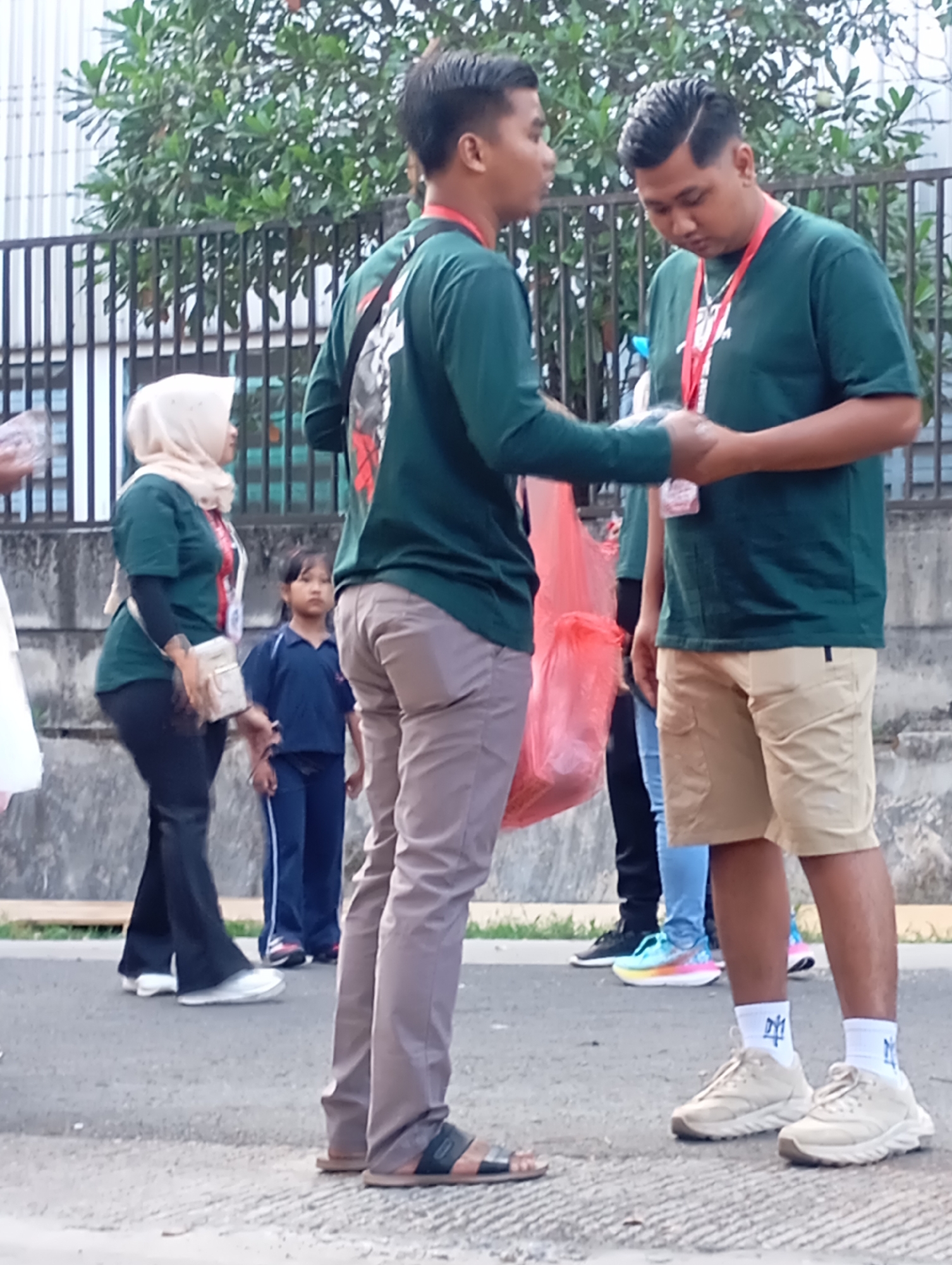 In Ngadimulyo, Indonesia, two young men in green t-shirts and lanyards interact on a paved street. The man on the left, wearing tan trousers, gestures with his hands while holding a red plastic bag. The man on the right, in beige shorts, looks down as they exchange an object. Behind them, a woman in a light-colored hijab and a green t-shirt walks away, followed by a young girl in a blue uniform. A black metal fence and green trees form the background.