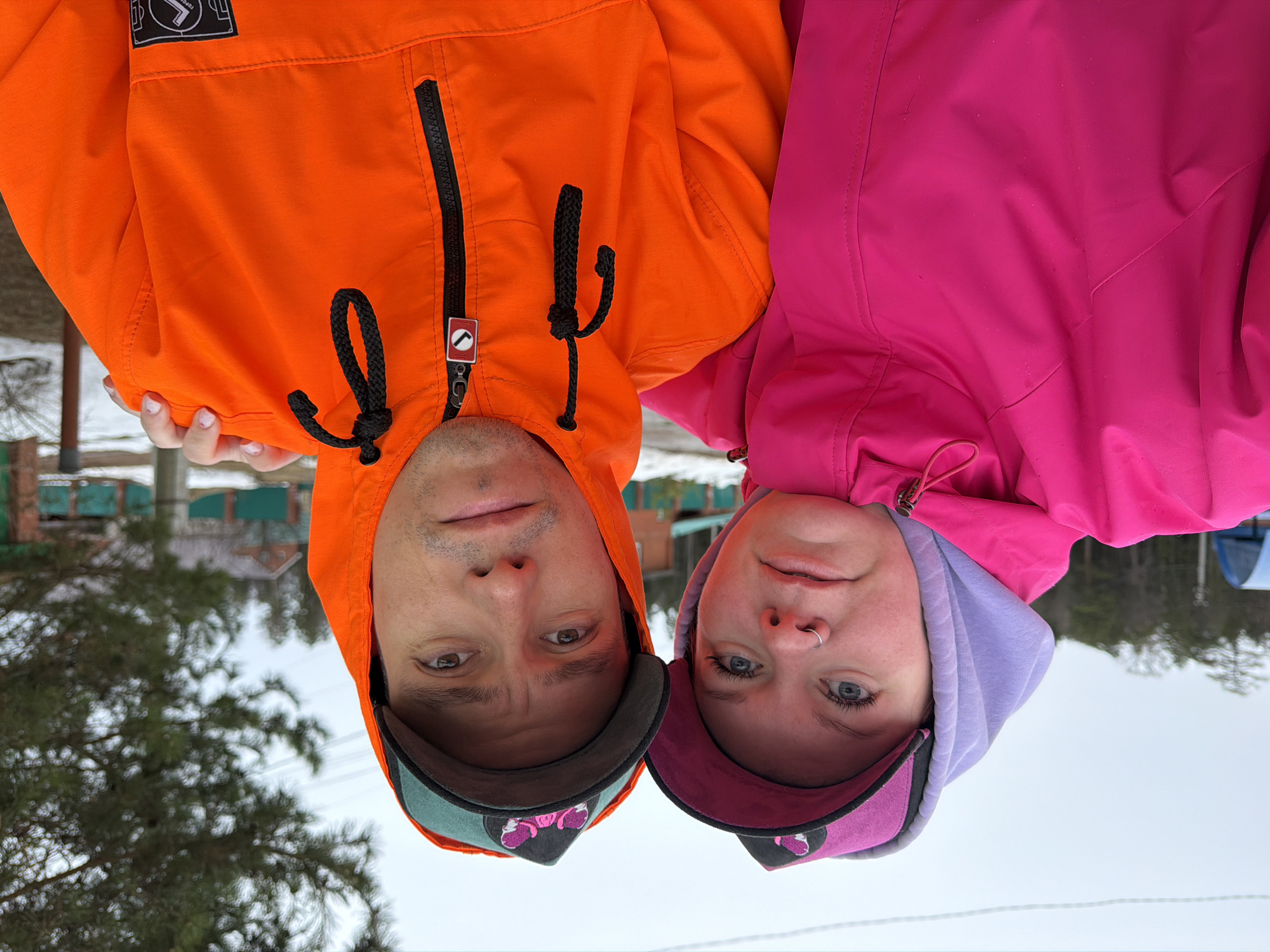 Two individuals are posing for a selfie in what appears to be an outdoor setting, likely during the colder months. The location may be Chaikovsky, Russia, though evidence within the image is not definitive.
The individual on the left is wearing an orange hooded jacket with a black zipper and black drawstrings. They are also wearing a baseball cap with a green brim. The individual on the right is wearing a pink hooded jacket with a lavender hood. They are also wearing a baseball cap with a pink brim. Both individuals are facing the camera with a neutral expression.
The background is somewhat blurred but appears to be a winter landscape with trees and possibly some buildings in the distance. The ground is covered with snow.