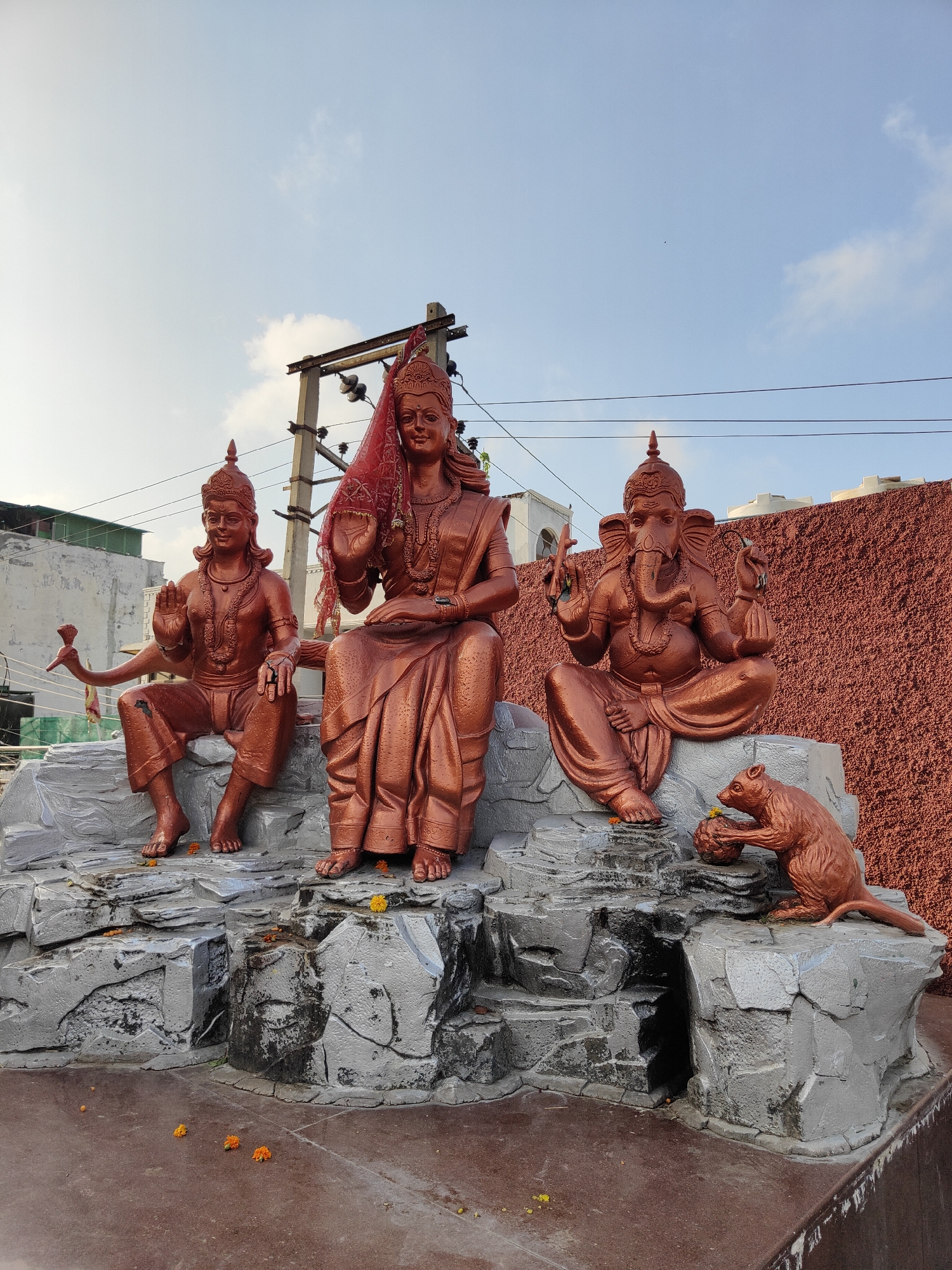 In Gurgaon, India, a vibrant outdoor scene unfolds featuring a religious shrine adorned with three bronze statues. The statues depict Hindu deities: Lord Vishnu on the left, Goddess Lakshmi in the center, and Lord Ganesha on the right. Lord Vishnu is portrayed seated, holding a bow and arrow, with a peacock fan behind him. Goddess Lakshmi is seated, draped in a red sari and veil, with a serene expression. Lord Ganesha, with his elephantine head, is also seated and appears to be in a meditative pose. A bronze statue of a mouse, Lord Ganesha's vahana, is positioned in front of him, holding a modak (a sweet dumpling). The statues are placed on a grey, rocky platform, which is decorated with scattered marigold flowers. The background features a textured reddish-brown wall and utility poles with electrical wires. The sky above is partly cloudy, indicating daytime. The overall ambiance suggests a place of worship or a cultural display.