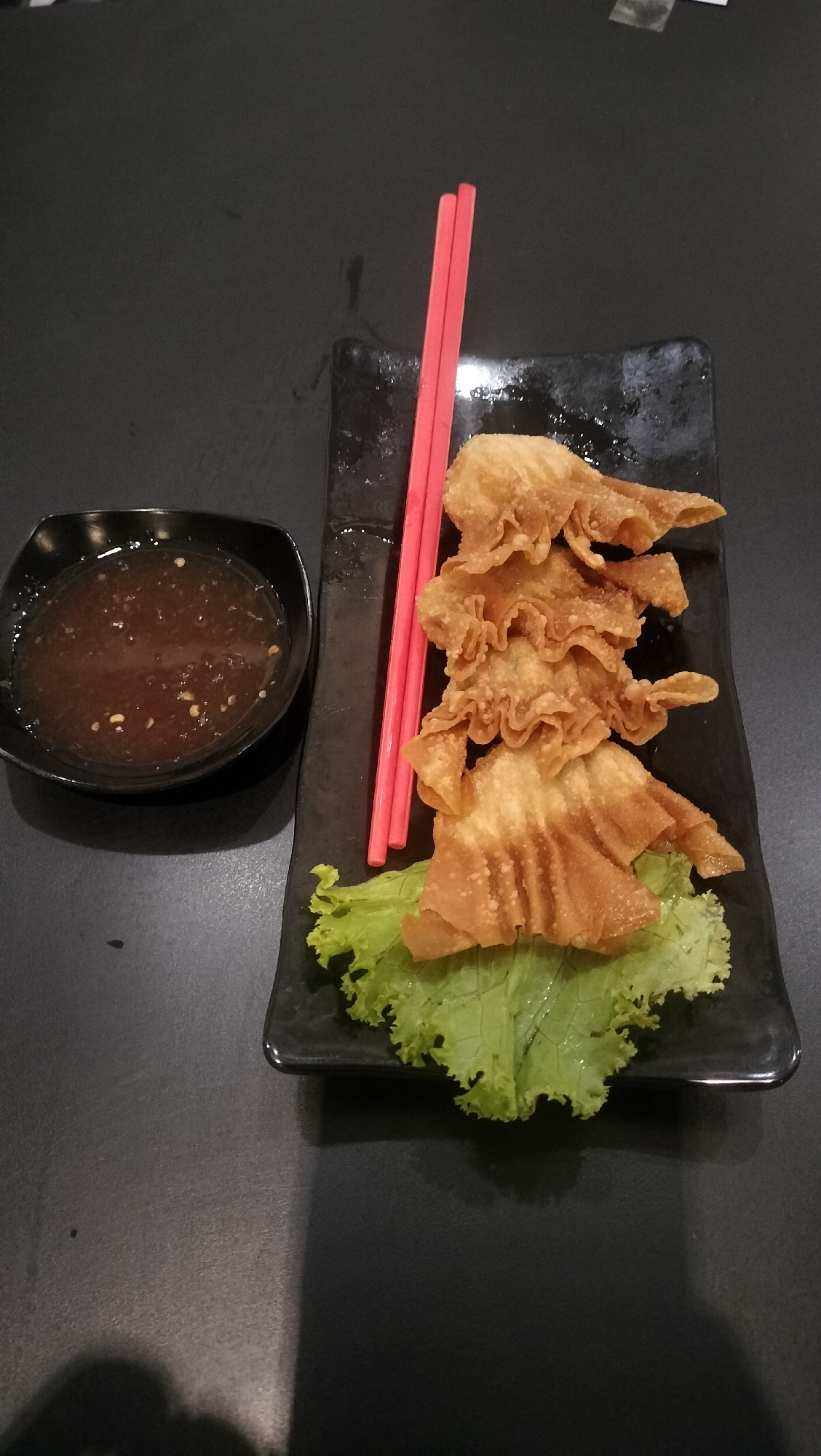 A high-angle medium shot shows a plate of fried wontons on a black surface. The plate is black and rectangular with rounded corners. Three crispy fried wontons are stacked vertically on a bed of green lettuce. Two red chopsticks are placed diagonally on the plate, resting across the wontons and lettuce. To the left of the plate is a small, black, square bowl filled with a dark, viscous dipping sauce containing visible flecks of what appear to be chilies or herbs. The background is a plain, dark, textured surface, likely a table. The lighting creates a slight shadow to the right of the plate. The scene is indoors, possibly in a restaurant.