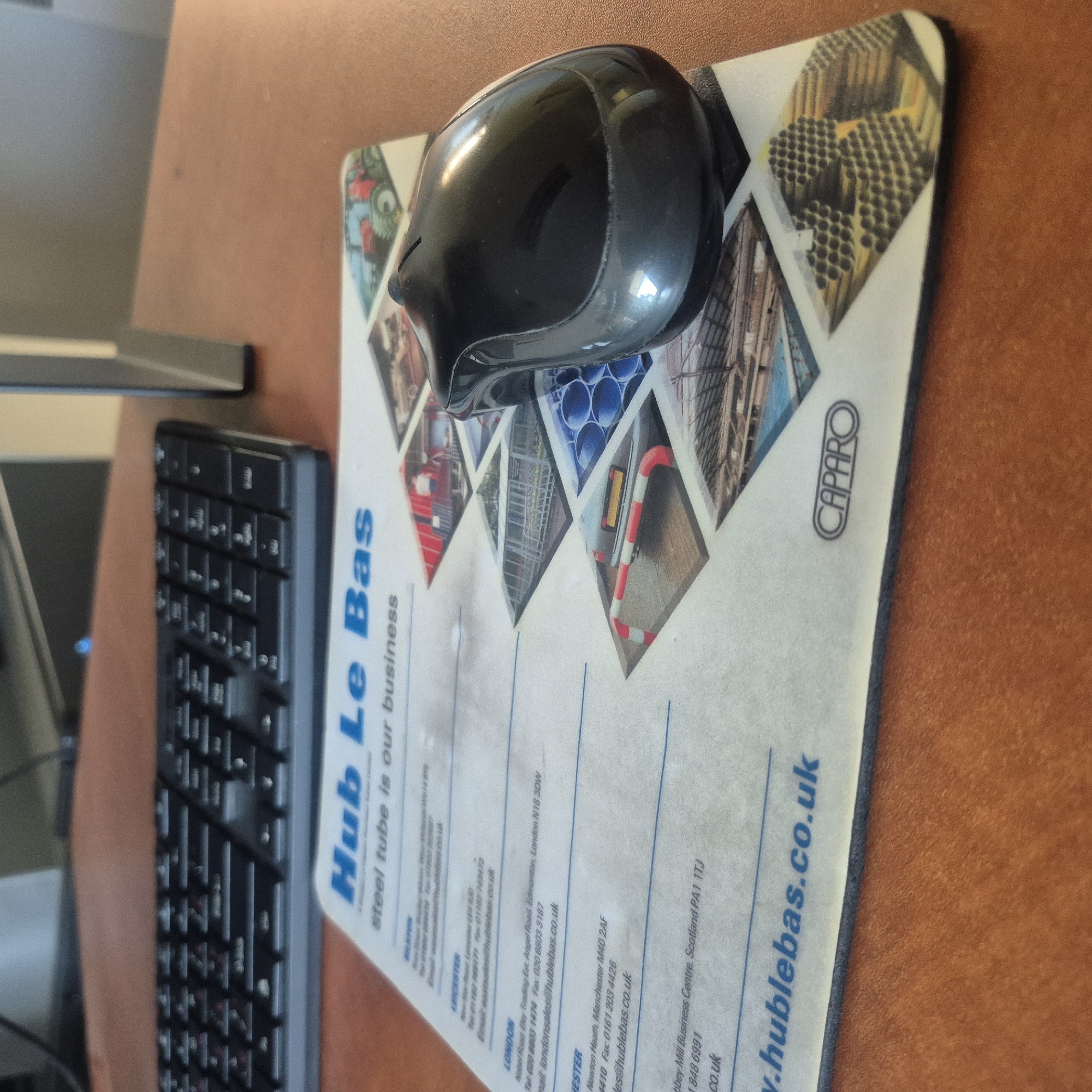 The image shows a desk with a black keyboard and a black computer mouse resting on a mousepad. The mousepad has a promotional design with multiple images of industrial materials and text. Visible text on the mousepad includes "Hub Le Bas", "steel tube is our business", "CAPARO", and website addresses like "www.hublebas.co.uk". There are also contact details for different locations such as "LEICESTER" and "LONDON", with phone numbers and email addresses. The mouse is glossy black and appears to be a standard optical or laser mouse. The keyboard is a compact, black keyboard with a numerical keypad on the right. The desk surface is a brown, wood-like material. The lighting suggests an indoor setting, likely an office or home workspace, with overhead lighting casting reflections on the mouse. There are no people visible in the image. The scene depicts a typical workspace setup.