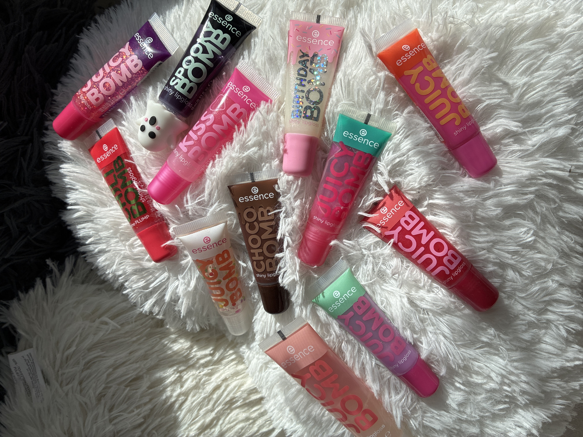 This media file features a top-down view of various lip gloss products from the brand "essence" scattered across a fluffy white rug. The lip gloss tubes are arranged in a scattered, somewhat haphazard manner, suggesting a casual display. The subjects are purely objects – multiple tubes of lip gloss, each with distinct colors and branding. One small white ghost figurine with black eyes is also visible among the lip gloss tubes.
The visible text on the lip gloss tubes indicates various product names and descriptions. Several tubes are labeled "JUICY BOMB" in large, bold lettering, with variations in tube color (purple, pink, green, orange, red, and a clear tube with pink lettering). Other visible product names include "SPOOK BOMB" (black tube), "KISS BOMB" (pink tube), "CHOCO BOMB" (brown tube), and "BIRTHDAY BOMB" (clear tube with pink and blue accents). The text also includes "essence" branding and "shiny lipgloss" descriptions, along with volume information like "10 ml/0.33 fl. oz." or "10m/0,33 fl. oz.". A small tag with text that appears to be "100% ΠΟΛΥΕΣΤΕΡΑΣPOLIESTER" is also partially visible at the bottom left.
The scene is set indoors, likely on a soft surface like a rug or blanket, indicated by the texture and the way the objects are settled. The lighting suggests it's daytime, with strong shadows cast by some of the lip gloss tubes, particularly on the left side of the image, hinting at sunlight coming from an angle. There is no discernible activity or event taking place; it appears to be a still life arrangement of beauty products. The overall mood is playful and colorful, due to the vibrant packaging of the lip glosses. The location context provided is Burgas, Bulgaria, but there are no visual cues in the image itself that specifically confirm this location.
