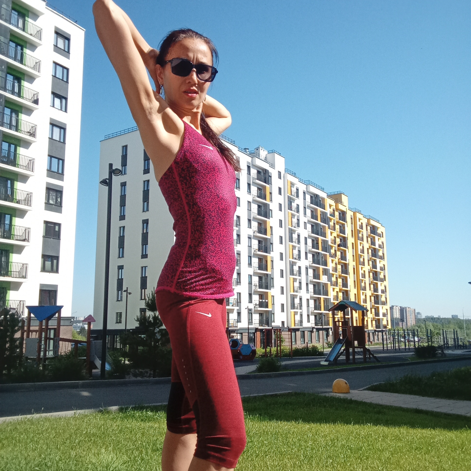 An adult female stands in a park with residential buildings in the background, under a clear blue sky. She is wearing a sleeveless, patterned pink athletic top and red capri-length athletic pants. Her hands are clasped behind her head, and she is wearing sunglasses. The environment includes a playground, a paved path, and a grassy lawn. The buildings appear to be apartment complexes. The location is the city of Pervomaysky, Russia.