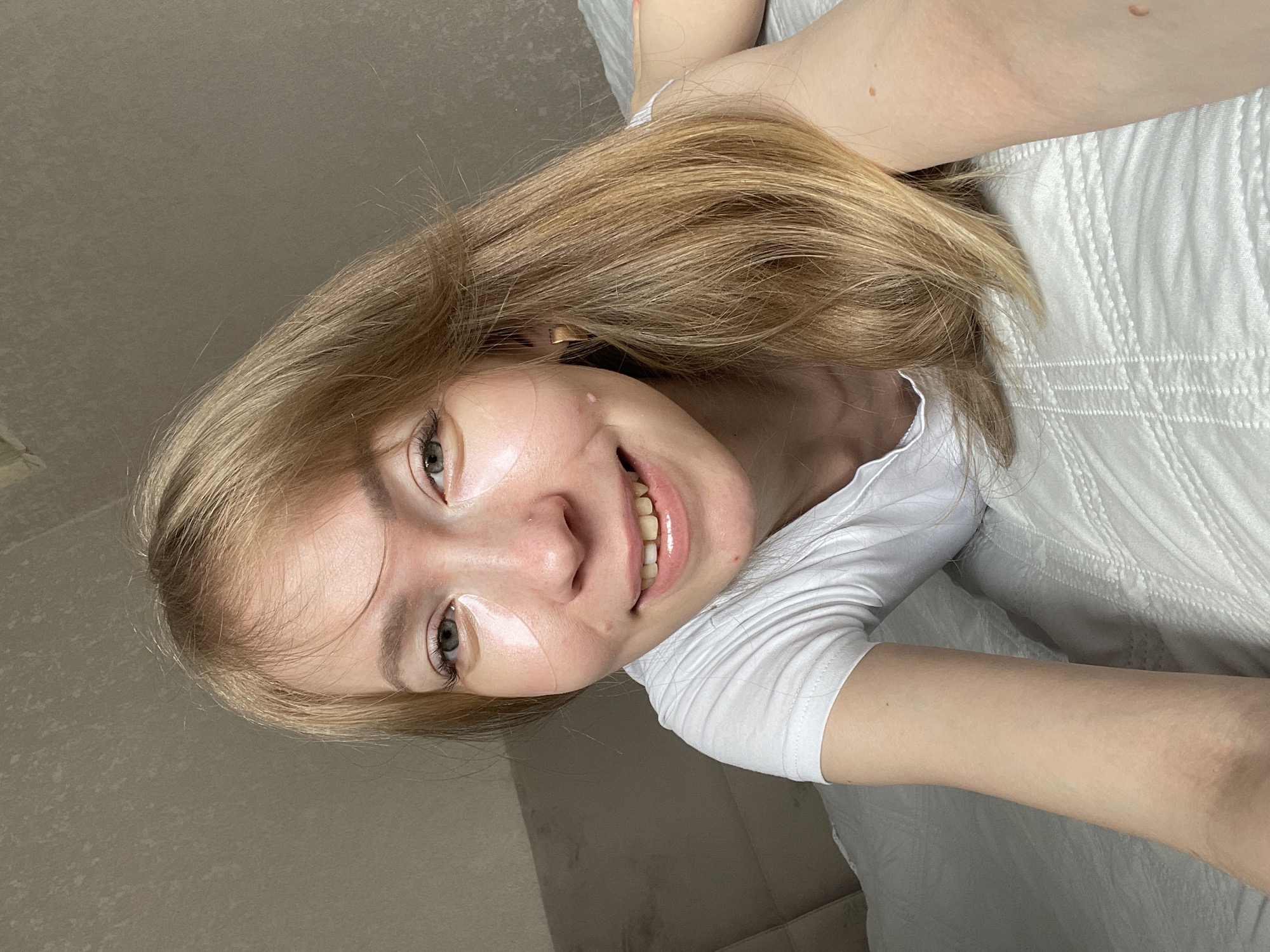 A fair-skinned young woman with light brown hair is lying on a bed, smiling at the camera. She is wearing a white t-shirt. A light-colored textured wall is visible in the background. The location in the city of Perm, Russia, cannot be determined from the image.