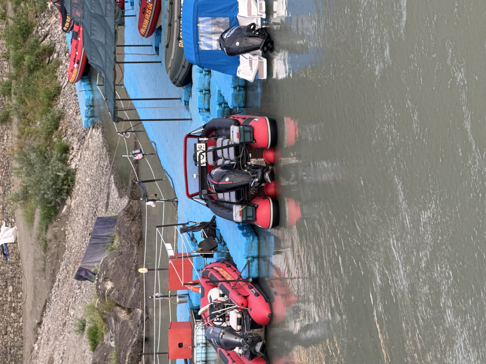 This image depicts a waterfront scene in the city of Barangol, Russia, likely at a docking area. Several inflatable boats are present, some red and some a dark green or gray color. One red inflatable boat is prominently positioned in the foreground, facing away from the viewer, and appears to be equipped with an outboard motor. To its left, another red inflatable boat, also with an outboard motor, is docked. In the background, more red inflatable boats are visible on blue floating platforms, and a boat covered with a blue tarp is docked. The water appears somewhat murky. The shoreline in the background is rocky with some vegetation. The overall impression is of a functional boating or rescue station. There are no people visible in the image.