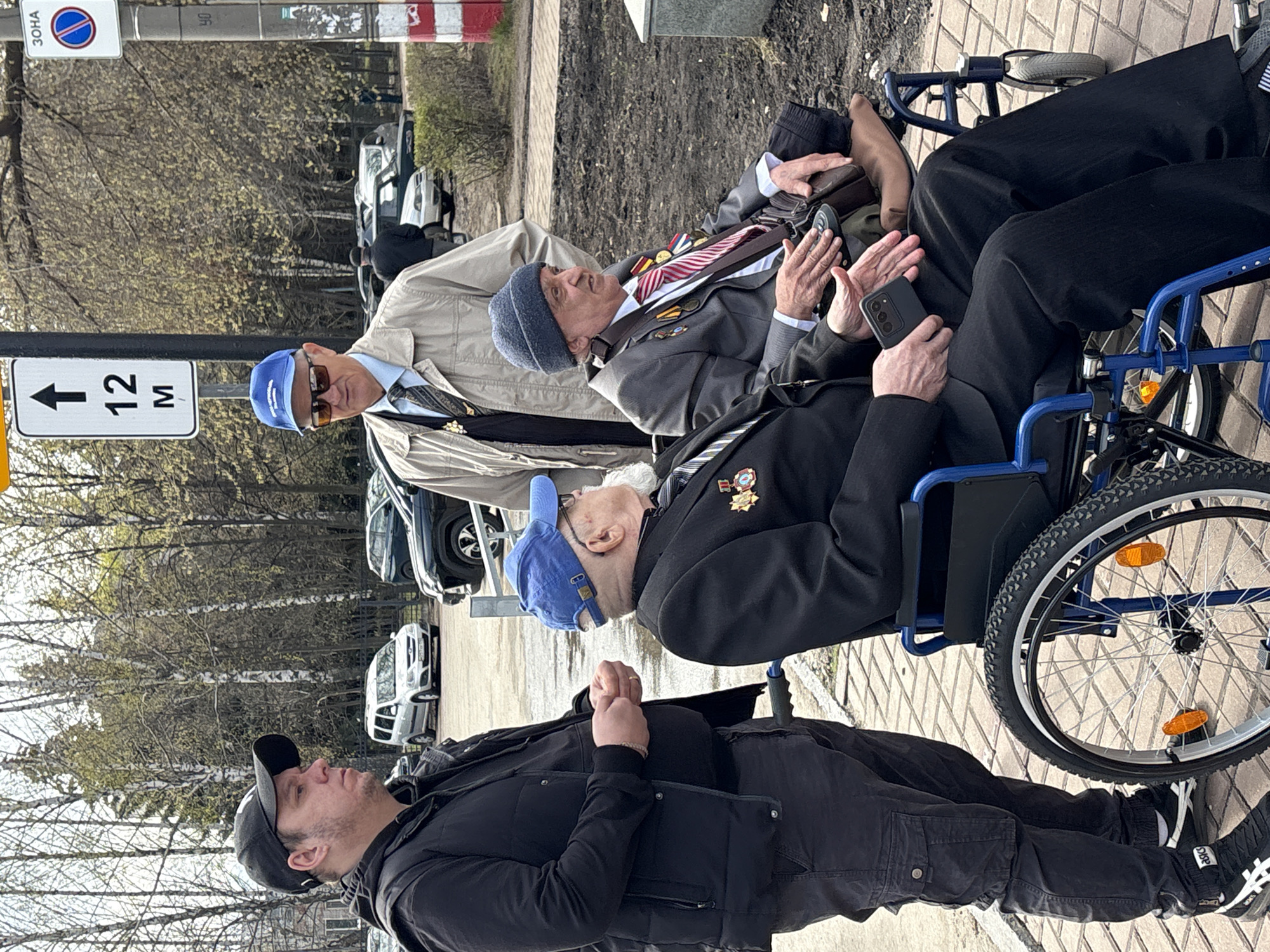 Three men stand near a street. Two of the men, one in a navy jacket and blue cap and another in a grey suit with medals and a striped tie and a knit cap, sit in wheelchairs. The third man, younger, wears a black jacket and cap. The man in the grey suit holds a smartphone. A street sign is partially visible, indicating "12 M" and a zone designation in Russian, characteristic of Ulyanovsk, Russia. Cars line the street in the background, indicating an urban environment. The curb is marked with red and white.