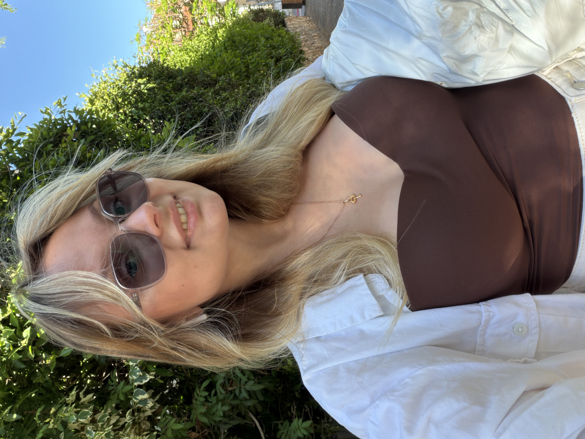 A medium shot depicts a fair-skinned young woman with long, blonde hair, angled slightly to the left, wearing sunglasses with a light-colored frame and tinted lenses. She is smiling and appears to be outdoors in Bat Yam, Israel. The woman is dressed in a dark brown top underneath an unbuttoned white shirt or jacket. A delicate gold necklace with a pendant is visible on her neck.
The background features lush green foliage, suggesting a garden or park setting. A glimpse of a clear blue sky is visible in the upper left corner. A building with a stone or brick facade is partially visible in the upper right background. The lighting suggests a sunny day.