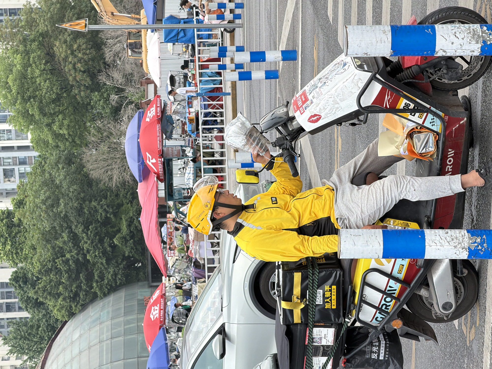 The image depicts an urban street scene in Chongqing High-tech Zone, China. In the foreground, a male delivery rider is seated on a red and yellow electric scooter. He wears a yellow long-sleeved uniform jacket, light gray sweatpants, black shoes, and a yellow helmet with a clear visor. The rider holds a clear plastic bottle, which appears to contain a foil-wrapped item, near the scooter's handlebars. A black delivery box with yellow straps, labeled with Chinese characters including "加入骑手 收入更高自由" (Join a rider, earn more freedom), is secured to the scooter's rear rack. A black plastic bag with white Chinese characters hangs from the rack. On the scooter's footrest, a light brown paper bag and another foil-wrapped item are present. A blue and white striped cylindrical bollard is partially visible in front of the scooter, with another positioned behind it, and more spaced along the adjacent street.
In the midground, a silver SUV is parked to the left of the scooter. Behind the scooter and the vehicle, an active open-air market is visible, featuring numerous stalls shaded by red, blue, and white umbrellas. Several individuals are present within the market area, some standing and others seated. A white metal barrier separates the market from the street.
The background displays a large glass dome structure on the far left. Dense green foliage dominates the central background, interspersed with some visibly bare trees. Taller, multi-story buildings are discernible in the distance beyond the trees. On the far right, a yellow construction excavator is partially visible behind market stalls and trees, alongside a yellow triangular road sign displaying a pedestrian crossing symbol mounted on a pole. The sky is overcast.