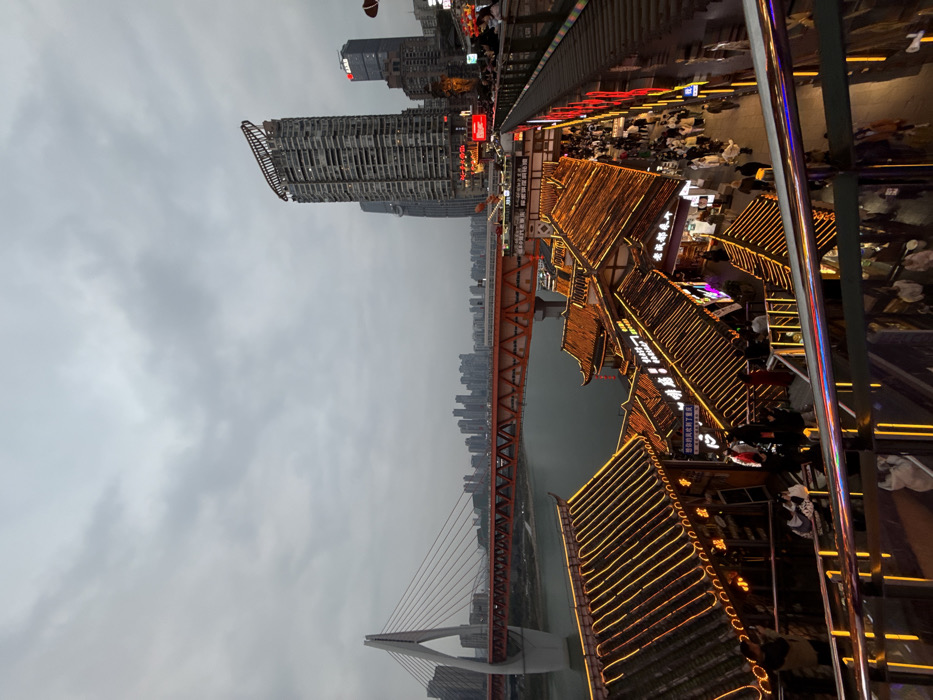 This image depicts a bustling, multi-layered urban scene at dusk in Yuzhong District, Chongqing, China, specifically showcasing the Hongyadong scenic area. The sky is overcast and grey.
In the foreground, an illuminated escalator or stairway with reflective surfaces leads downwards. Below and extending into the midground are numerous traditional Chinese-style buildings with multi-tiered, dark tiled roofs, extensively outlined with golden LED strip lights. These structures house shops and restaurants, indicated by various illuminated signs with Chinese characters. A large number of pedestrians are visible on the walkways and platforms between these buildings, moving about and observing the surroundings.
To the left of the traditional buildings, the dark waters of the Jialing River flow. Spanning the river are two prominent bridges. The primary bridge is a white cable-stayed structure with reddish-orange suspension cables and a white pylon, extending towards a distant cityscape. Parallel to and slightly behind this bridge is a reddish-orange truss bridge, possibly carrying a railway, with a train-like object visible on its deck.
On the right side of the image, towering above the traditional architecture, stands a modern, tall skyscraper with a distinctive curved, open-frame structure at its apex. Adjacent to it are other high-rise buildings, one displaying an illuminated "BANK" sign. Further in the background, a dense urban skyline of additional high-rise buildings is visible against the grey sky, extending across the river. The overall environment is characterized by a blend of traditional and modern architecture, significant infrastructure, and active pedestrian life under artificial illumination.