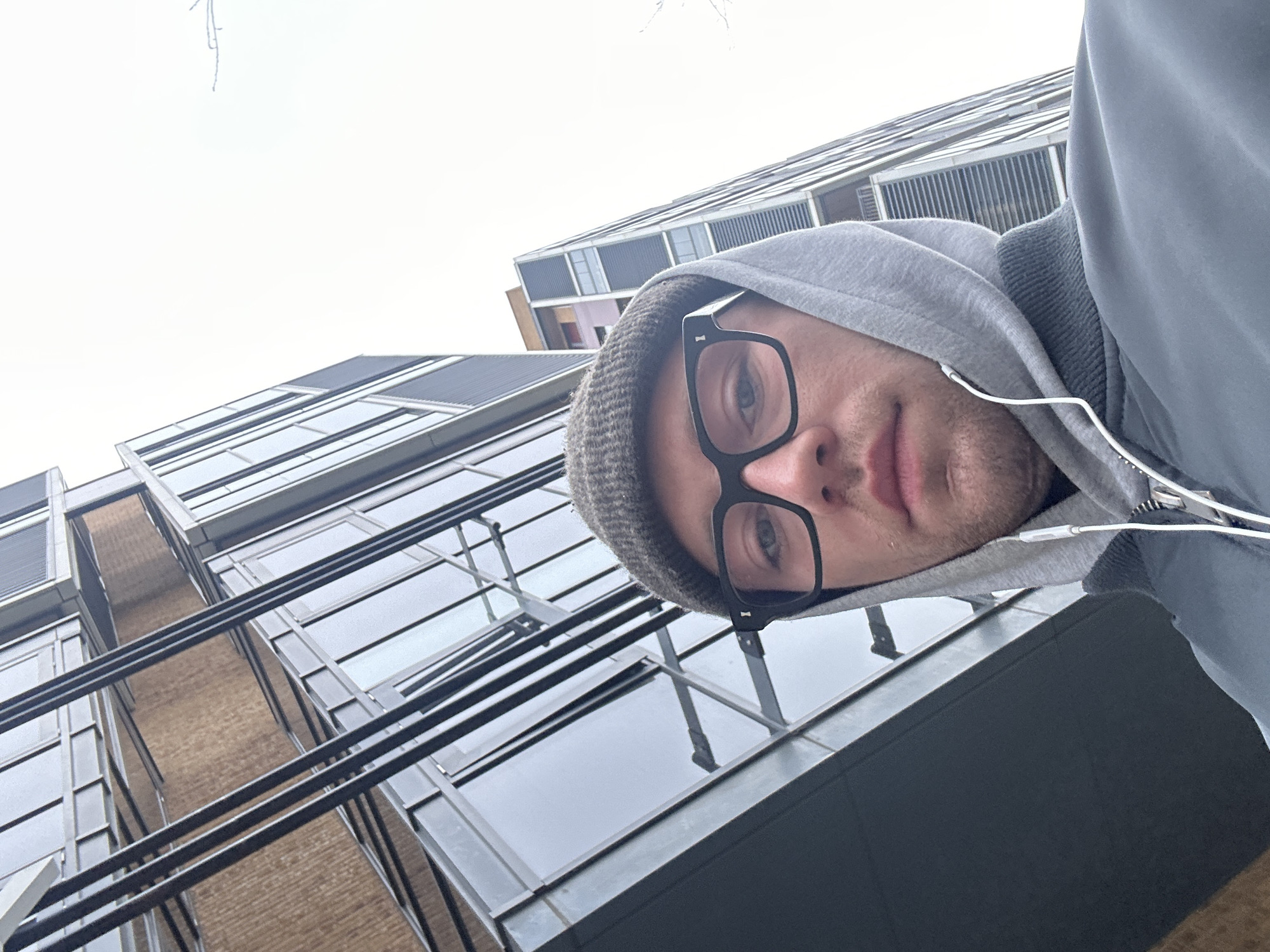 A low-angle shot shows a person wearing glasses, a beanie, and a grey hoodie with earbuds, positioned against a backdrop of modern architecture in London, United Kingdom. The buildings feature dark-framed glass windows and metallic structures. The sky appears overcast.