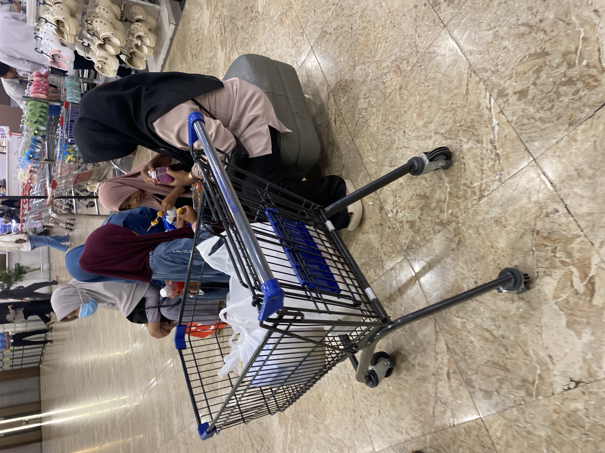 In a commercial setting with marble flooring, a shopping cart filled with white plastic bags is prominently featured in the foreground. Behind the cart, a group of individuals, predominantly women and children wearing hijabs, are seated. One woman, wearing a light grey hijab and a light blue face mask, is partially visible. Another woman in a dark maroon hijab is holding a red bottle. A child in a pink hijab is holding a skewer with food and a small purple bottle. A woman in a black hijab and a light pink top is seated on a dark grey ottoman. The background shows shelves stocked with colorful flip-flops and other merchandise, as well as other shopping carts. The overall scene suggests a family shopping trip in a store, possibly a department store or supermarket, in Banda Aceh, Indonesia.