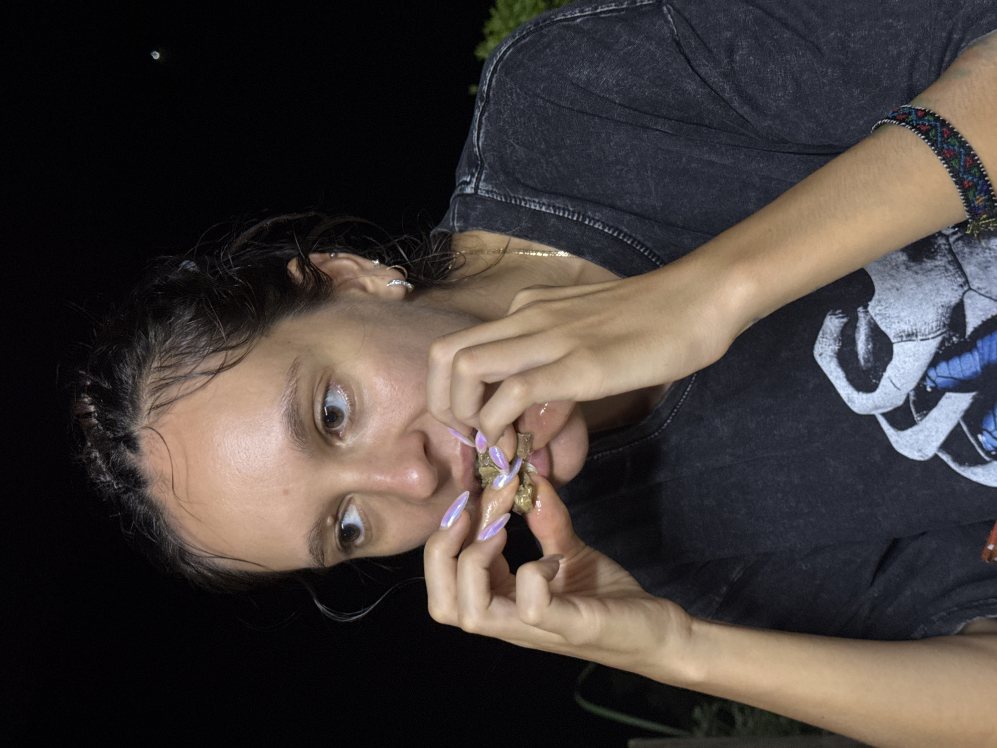 A woman with light skin and dark hair is eating something with her hands. She has iridescent, pointed nails. She is wearing a gray t-shirt with a white skull graphic and a colorful bracelet. The background is dark, suggesting it is nighttime. The location appears to be outdoors. There is no indication it is in Pờ Ly Ngài Commune, Vietnam.