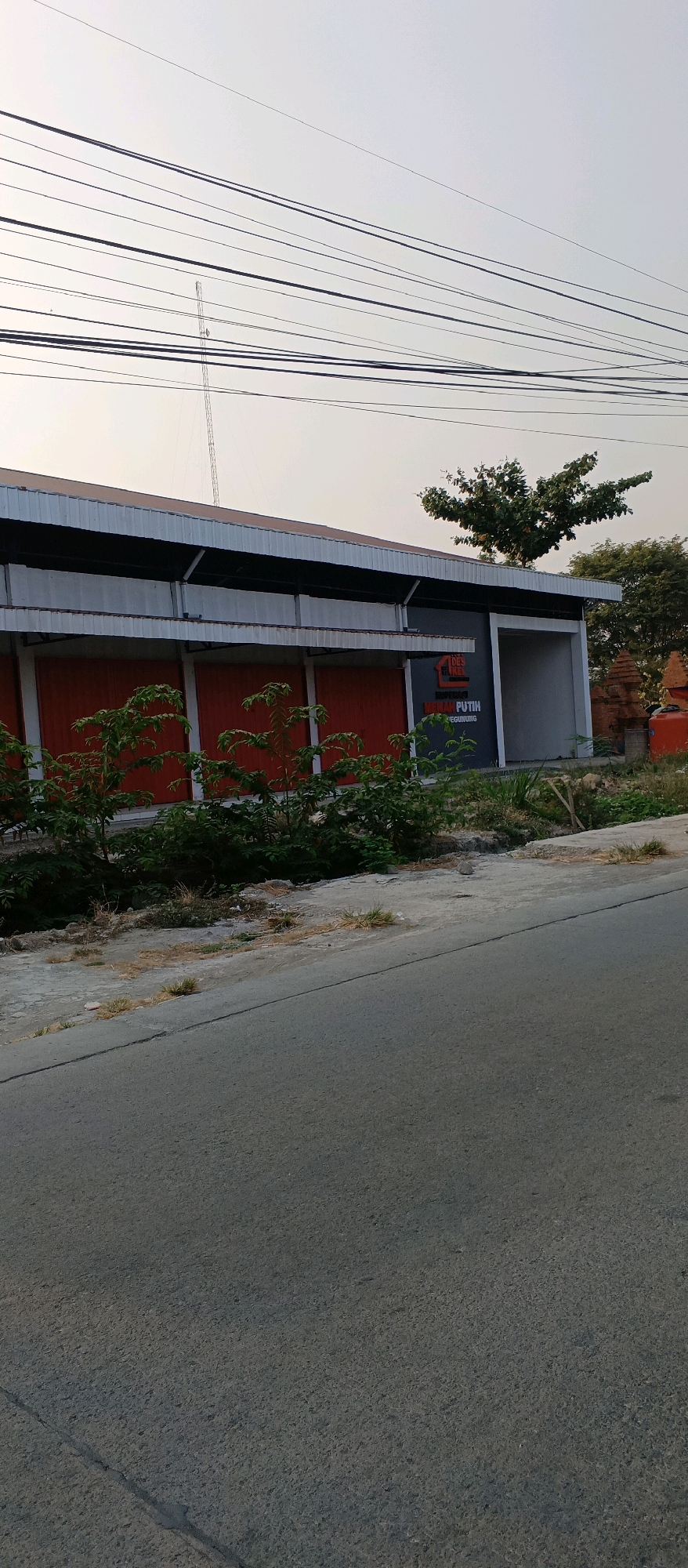 The image captures a storefront in the city of Gegunung, Indonesia. The building features a metallic roof and red roll-up doors, with a partial view of a sign that reads "DES KEMILAU PUTIH." Green foliage is visible in front of the building, and power lines crisscross the sky above. The foreground shows a grey asphalt road. The scene is illuminated by natural light, suggesting it was taken during the daytime.