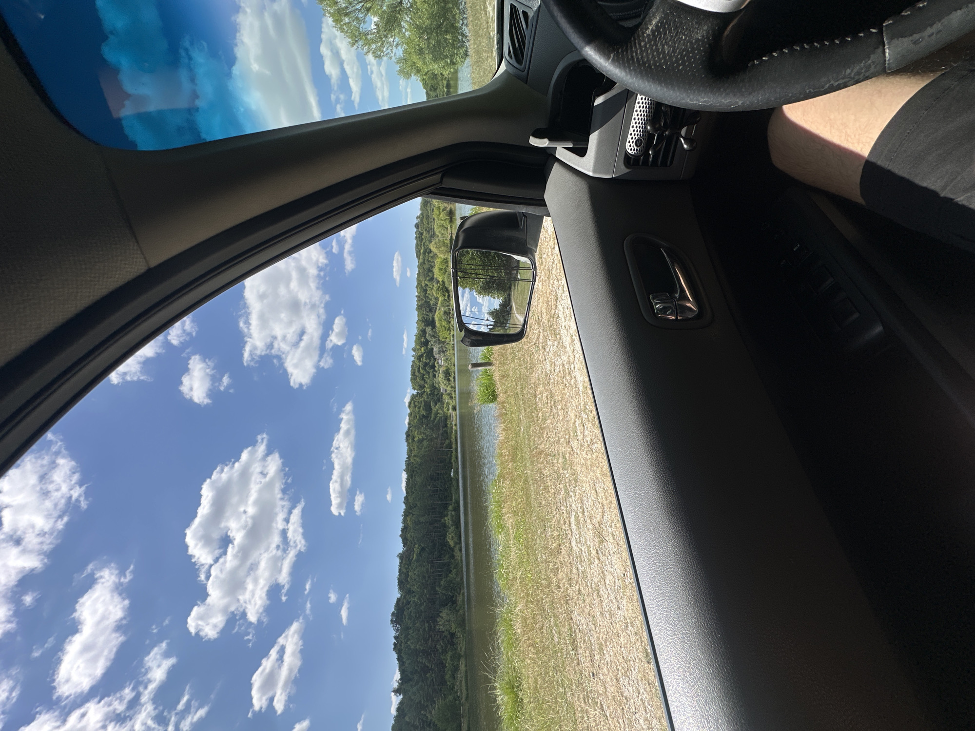 The image is taken from inside a car, looking out the passenger window and side mirror. The scene depicts a sunny day with a bright blue sky filled with fluffy white clouds. A body of water, likely a lake or river, is visible in the mid-ground, bordered by a line of trees on the far bank. The foreground shows dry, grassy terrain and the interior of the car, including the door panel, a side mirror, and part of the steering wheel and a person's leg. The reflection in the side mirror shows trees and a pathway. While the image depicts a natural landscape, there is no indication of the specific location within the city of Makiv, Ukraine.