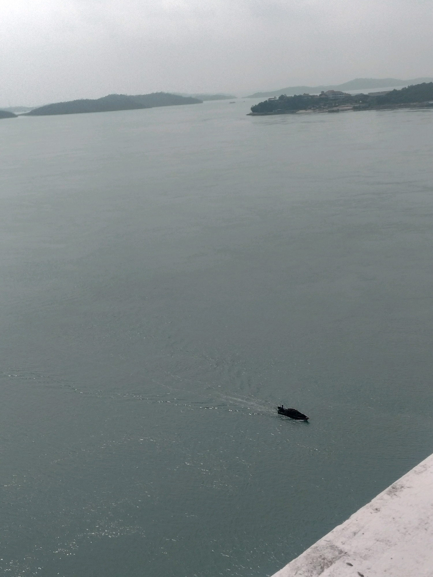 The image depicts a wide expanse of calm, grey water with several islands dotting the horizon. In the foreground, a white railing or structure is partially visible, suggesting the viewpoint is from a bridge or elevated platform. A solitary, dark-colored boat is navigating the water, leaving a faint wake behind it. The islands in the background are covered in dense greenery, with some structures visible on one of the larger islands, hinting at human habitation or development. The sky is overcast, contributing to the muted color palette of the scene. The overall impression is one of a tranquil, perhaps slightly hazy, day in a coastal or island region. This scene is characteristic of many locations around Batam, Indonesia, particularly areas with island chains and waterways like the one connecting Batam Island to its neighboring islands, such as the waters near Nongsa or Sekupang.