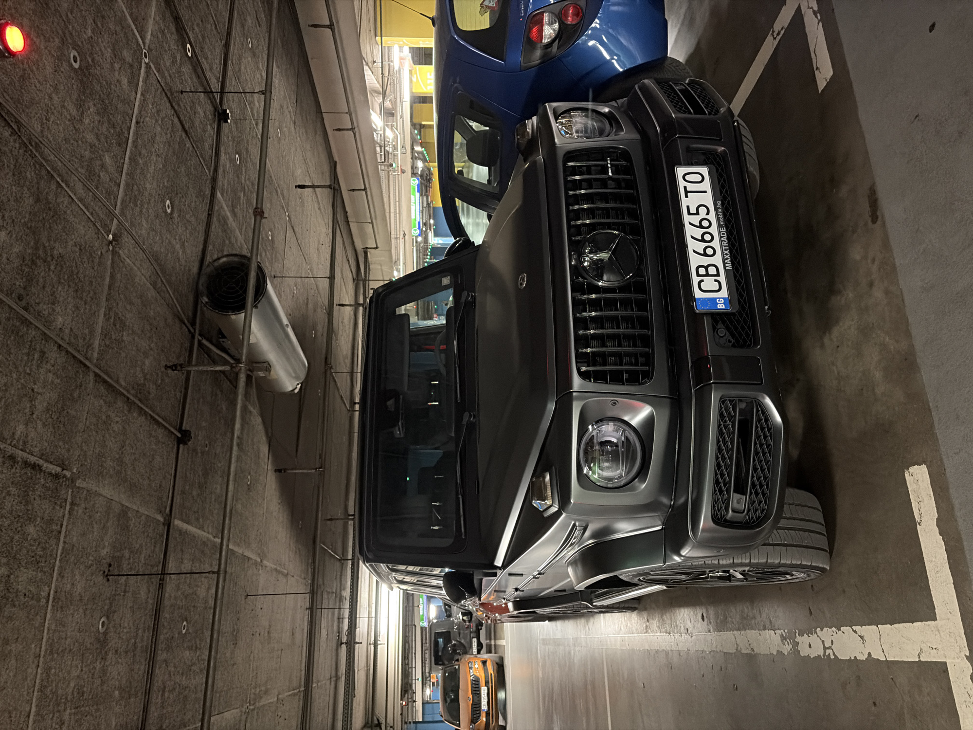 An exterior, eye-level, medium shot captures a matte black Mercedes-Benz G-Class SUV parked in an underground parking garage. The vehicle is angled slightly towards the camera, with its front grille, headlights, and license plate prominently displayed. The license plate reads "CB 6665 TO" and has the "BG" abbreviation for Bulgaria. The vehicle is in a parking space marked by white lines on the concrete floor. In the background, other vehicles are visible, including an orange SUV and a blue hatchback, along with concrete pillars and a yellow-painted wall. The ceiling of the garage features exposed pipes, ventilation systems, and lighting fixtures. A red light is visible in the upper right corner of the image. The scene suggests a common urban environment, likely within the city of Sofia, Bulgaria, given the license plate information. No people are visible in the image.