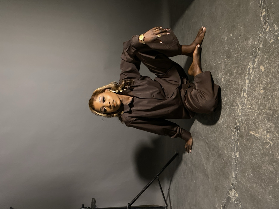 The image captures an adult female posing in a photography studio in Jos, Nigeria. She is seated on a dark grey concrete floor, which shows some subtle scuff marks, against a plain, lighter grey seamless backdrop. A black metal stand, likely for lighting equipment, is partially visible on the far left.
The subject has a dark complexion and light brown hair with blonde highlights, styled in loose waves, falling over her shoulders. She wears prominent makeup, including defined eyebrows, winged eyeliner, and a glossy nude lip color. Her accessories include long, multi-tiered gold-toned dangling earrings, a gold-toned wristwatch on her left wrist, and several gold-toned rings on her fingers. Her fingernails are painted with a light, neutral polish and are long.
She is dressed in a matching two-piece ensemble consisting of a dark brown, long-sleeved button-up shirt and dark brown wide-leg trousers. She is barefoot. Her body is oriented towards the right side of the frame, with her torso slightly turned. Her left leg is extended forward, resting on the heel, while her right leg is bent at the knee, with the foot tucked in towards her body. Her left arm is extended back, with her hand resting flat on the floor for support. Her right arm is bent, with her hand resting on her right knee. Her gaze is directed slightly off-camera to her left, with a composed expression.