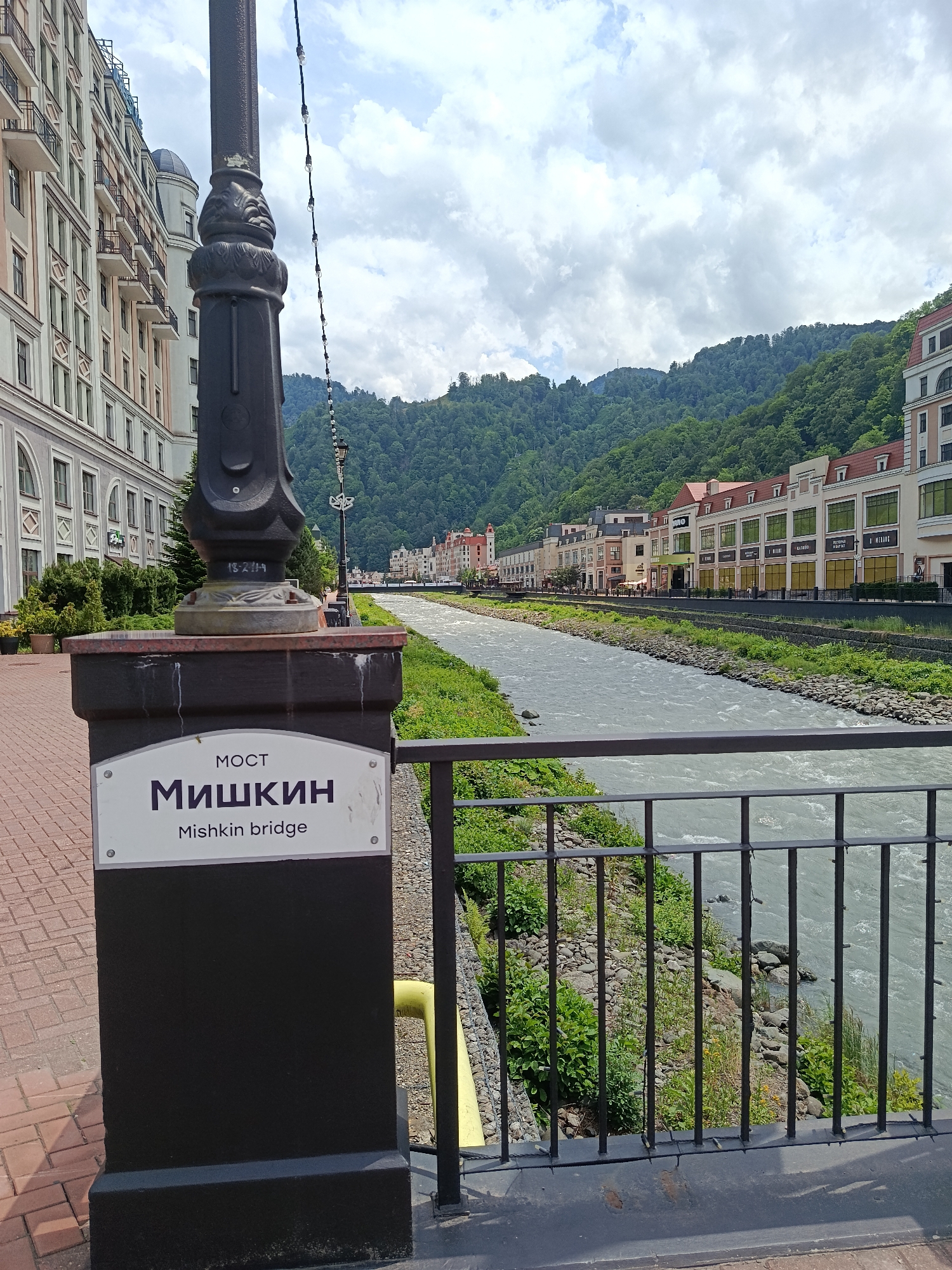 This media file depicts a scenic view of a river flowing through a town nestled in a mountainous region. In the foreground, a black lamppost stands prominently, adorned with a decorative base and a string of lights. Attached to the base is a sign with text in both Russian and English, reading "МОСТ Мишкин Mishkin bridge". The bridge itself is implied by the sign and the railing that stretches across the image, overlooking the river.
The river is wide and appears to be flowing rapidly, with rocky banks lined with lush green vegetation. On the far side of the river, a row of multi-story buildings with red-tiled roofs stretches along the bank, creating a charming architectural facade. Behind these buildings, steep, densely forested mountains rise, their slopes covered in vibrant green trees. The sky above is partly cloudy, suggesting a pleasant, possibly warm day.
The overall scene evokes a sense of tranquility and natural beauty, with the man-made structures seamlessly integrated into the mountainous landscape. The location appears to be a resort town, likely in a place like Estosadok, Russia, given the presence of resort-style architecture and the mountainous environment. No people are visible in the image, emphasizing the peaceful atmosphere of the location. The time of day seems to be daytime, given the lighting and cloud cover.