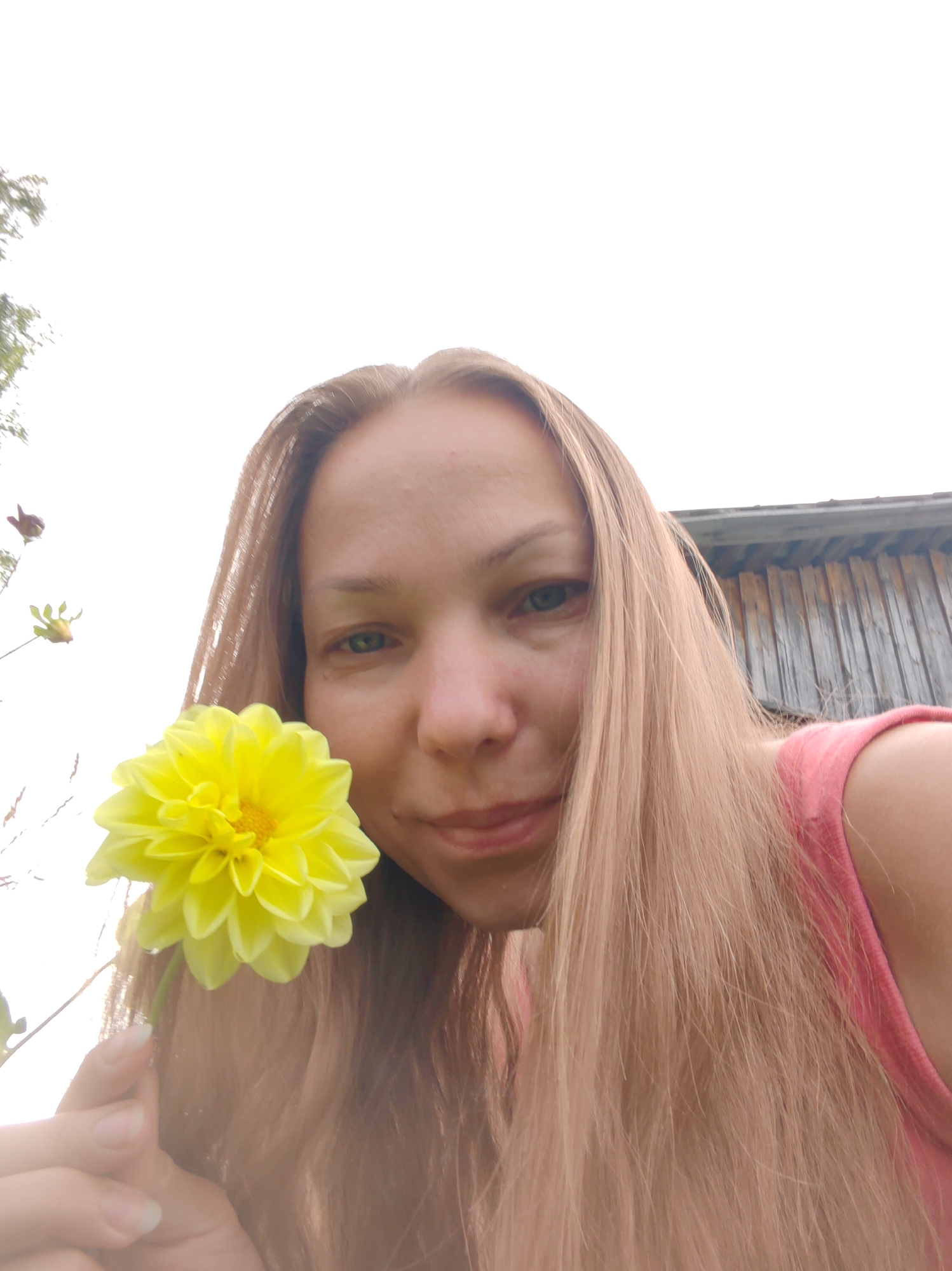 A woman, with long blonde hair and fair skin, is in the foreground of this image. She is looking towards the camera, with a slight smile. She is holding a bright yellow flower in her right hand. Behind her, to the right, is a weathered wooden building. To the left of the woman, there are some branches with a few more flowers or buds. The sky is overcast and bright. The image appears to be taken outdoors, likely in a rural or garden setting in the city of Chukhlem, Russia.