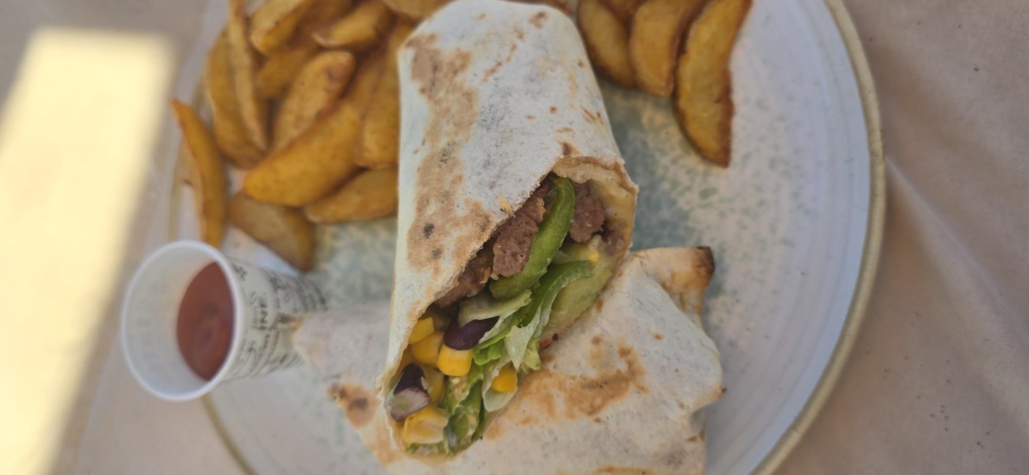 The image shows a plate of food featuring a cut burrito, potato wedges, and a small cup of sauce. The burrito contains ingredients such as lettuce, corn, beans, meat, and green pepper. The potato wedges are golden brown and appear to be seasoned. The sauce in the cup is red. The plate is light-colored with a speckled pattern and a thin, light-colored rim. The food is placed on a beige surface with some dark specks. The scene appears to be set outdoors.