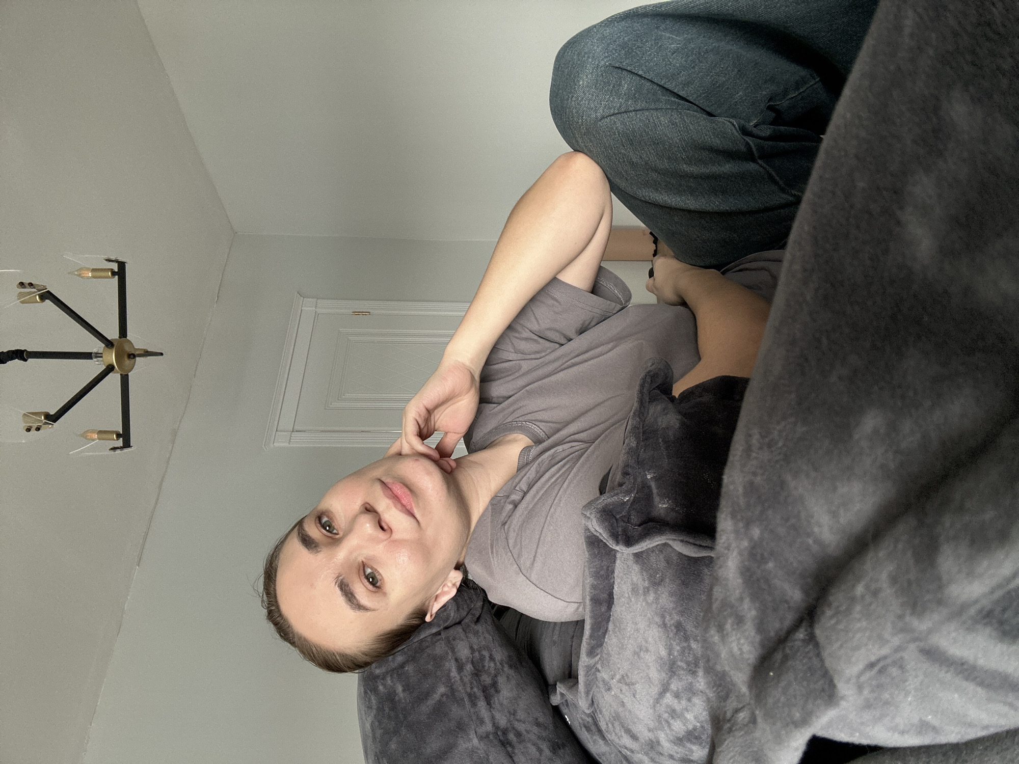 A woman lies on a gray plush blanket, with her head resting on the back of what appears to be a gray upholstered couch. She wears a gray t-shirt, and her left hand is raised to her face, with her fingers gently touching her cheek. The woman gazes towards the camera. Above her, a black and gold modern chandelier is affixed to the ceiling. In the background, a white door is partially visible. The bottom right of the frame is heavily obscured by more of the gray blanket. In the upper-right, a portion of a person's blue jeans clad legs are visible. There is no verifiable information regarding the location being in Kharnavy, Russia.