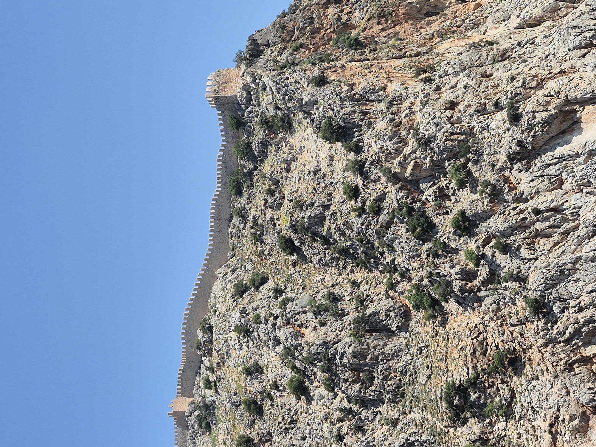 The image shows a defensive stone wall, likely part of the Alanya Castle in Alanya, Turkey, built along the side of a steep, rocky cliff face. The wall has crenellations along its top edge. Sparse vegetation, consisting of green shrubs and trees, is scattered along the rock face. The sky in the background is clear and blue. The camera is angled upwards, emphasizing the height and steepness of the wall and cliff.