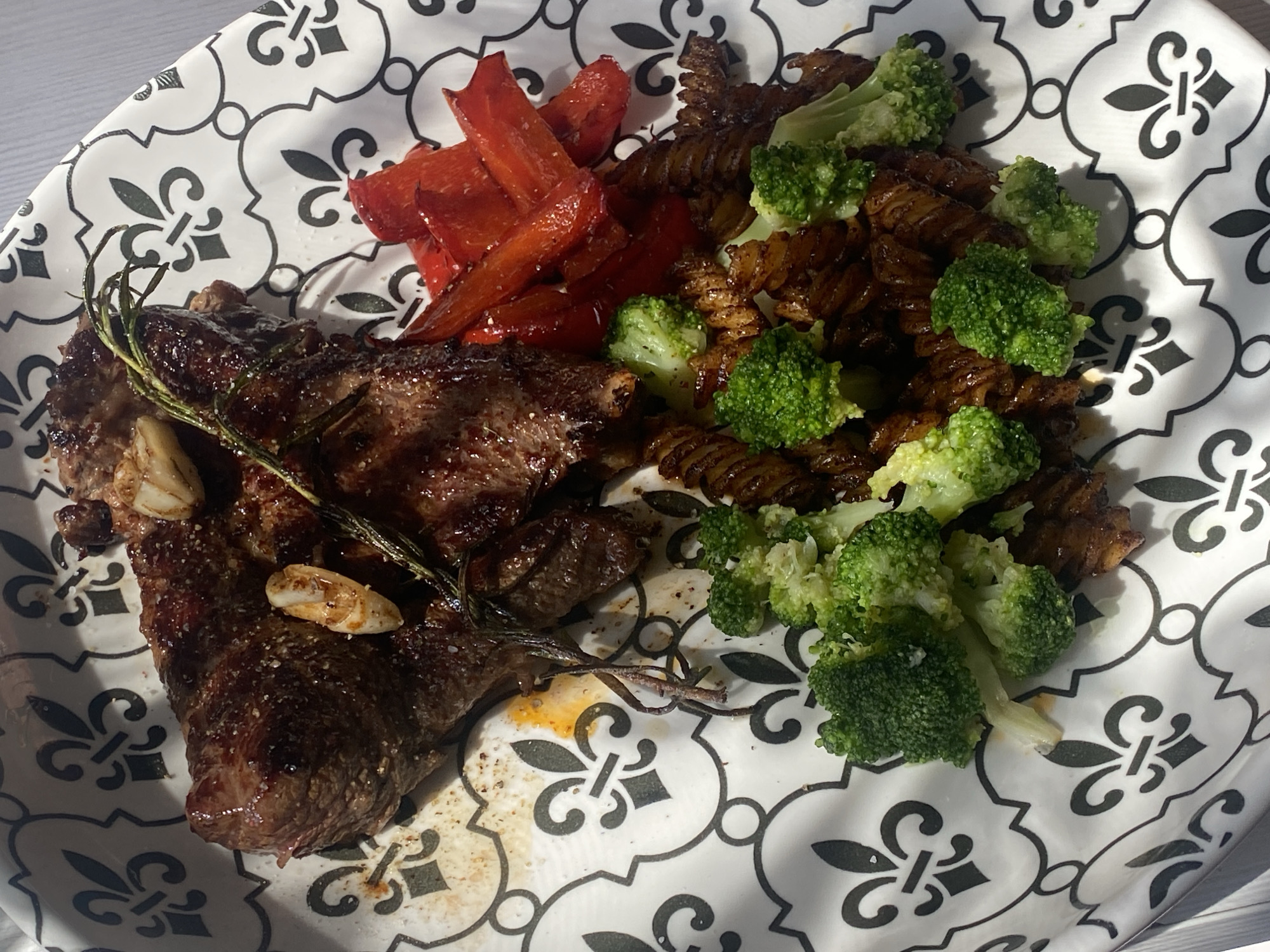 A close-up, high-angle shot captures a prepared meal served on a decorative white plate with a repeating black ornamental pattern, possibly a stylized fleur-de-lis motif. The meal features a seared steak, garnished with fresh rosemary sprigs and roasted garlic cloves, exhibiting a brown crust and visible seasoning.
Alongside the steak are bright red bell pepper strips, likely roasted or sautéed. On the opposite side of the plate, dark spiral pasta (fusilli) is mixed with numerous vibrant green broccoli florets. The pasta appears to be coated in a dark sauce.
The scene is brightly lit by natural light, casting distinct shadows across the food and plate, suggesting daytime, possibly an afternoon setting. No people are visible, and there is no discernible text within the image. The context indicates the location is Chaikovsky, Russia, though no specific visual cues confirm this. The food is presented neatly, suggesting it is ready for consumption.