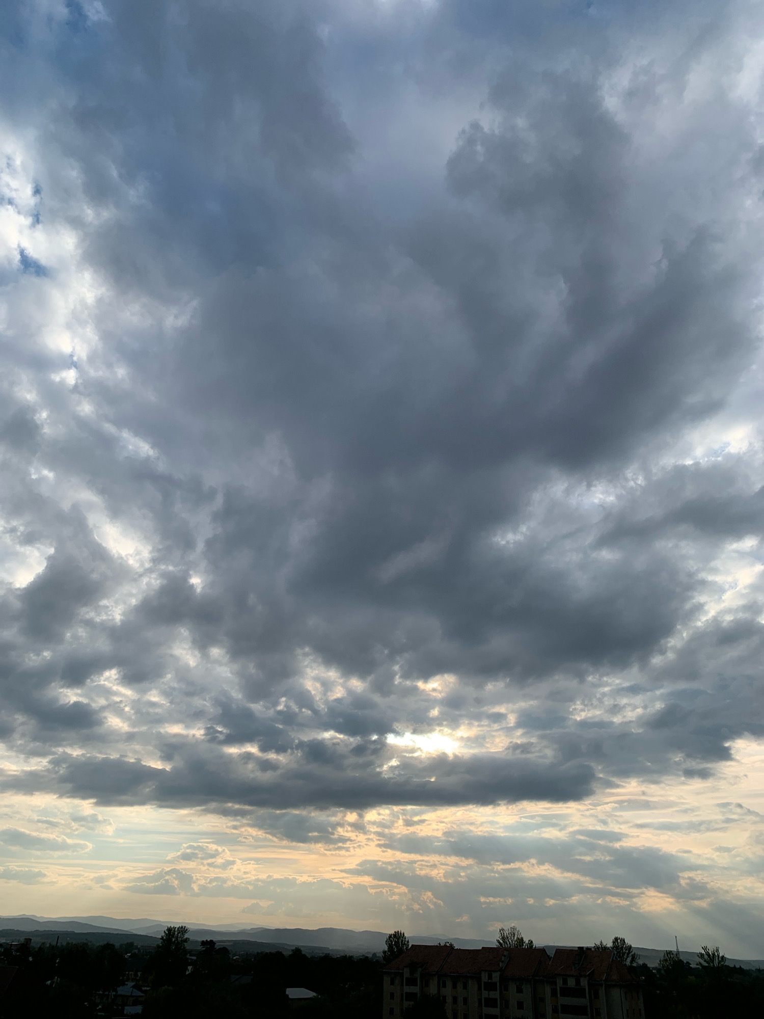 This image captures a dramatic sky over the city of Kolomyia, Ukraine, likely taken from an elevated viewpoint. The sky is filled with a dense layer of dark, brooding clouds, suggesting an impending storm or a recent downpour. Shafts of sunlight pierce through gaps in the clouds, creating dramatic crepuscular rays that illuminate the landscape below.
In the foreground, silhouetted against the bright sky, are the dark shapes of trees and the rooftops of buildings. The architecture appears to be residential, with several multi-story structures visible. The terrain is rolling, with hills and mountains forming a distant horizon line. The overall impression is one of natural power and the beauty of a dynamic sky. There are no people or specific actions depicted in the image. The environment is natural and urban, with a focus on the atmospheric conditions.