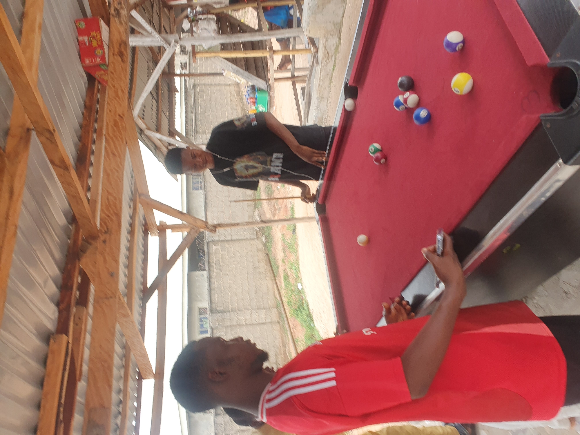 In Lokoja, Nigeria, two young men are engaged in a game of pool under an open-air shelter with a corrugated metal roof supported by wooden beams. The man on the left, wearing a red shirt with white stripes and black pants, stands on the left side of a red pool table and is focused on the game. He is holding a cue stick in his right hand and appears to be lining up a shot. On the right side of the pool table, another young man in a black t-shirt with a graphic print and black shorts is actively playing, holding a cue stick and about to strike a white cue ball. Several pool balls, including a white cue ball, a red and green striped ball, a solid red ball, a solid blue ball, a yellow ball, a purple ball, and a black ball, are scattered across the red felt surface of the pool table. The environment is informal, suggesting a recreational area.