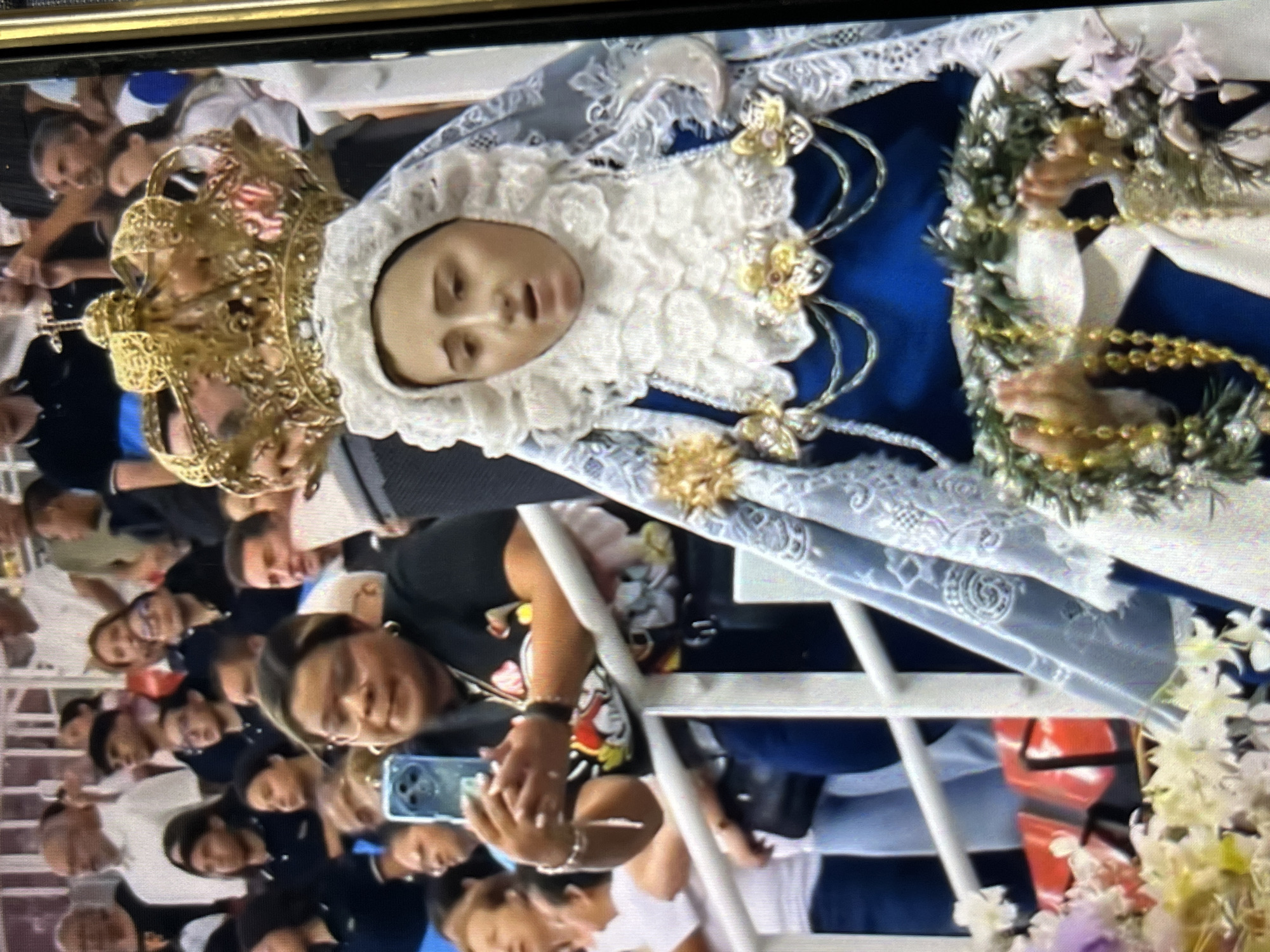 A photo shows a religious procession in Tinaquillo, Venezuela. The focal point is an effigy of a saint, crowned and ornately dressed in blue and white. The effigy is surrounded by people, some of whom are taking photos with their phones. The environment appears to be an outdoor public space, possibly during a festival or religious celebration.