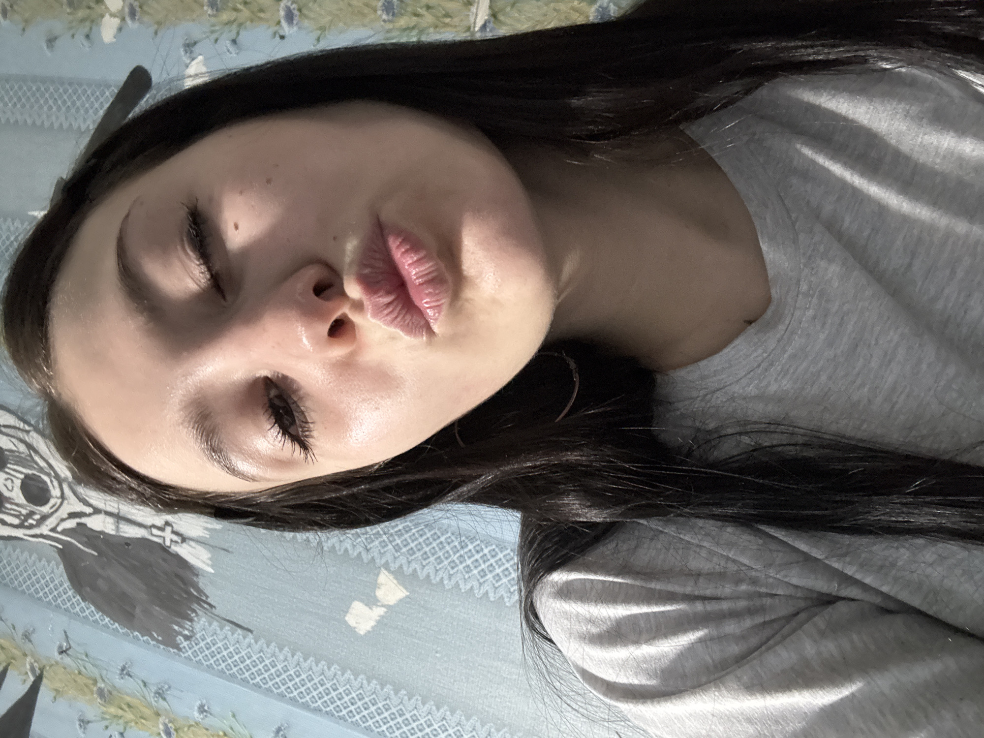 The image is a close-up selfie of a young woman with dark hair. She is lying down, facing the camera with her eyes partially closed and her lips pursed in a kiss. She is wearing a light gray shirt. The background consists of a blue patterned fabric. There is no information in the image indicating that the location is in the city of Фоки, Russia.