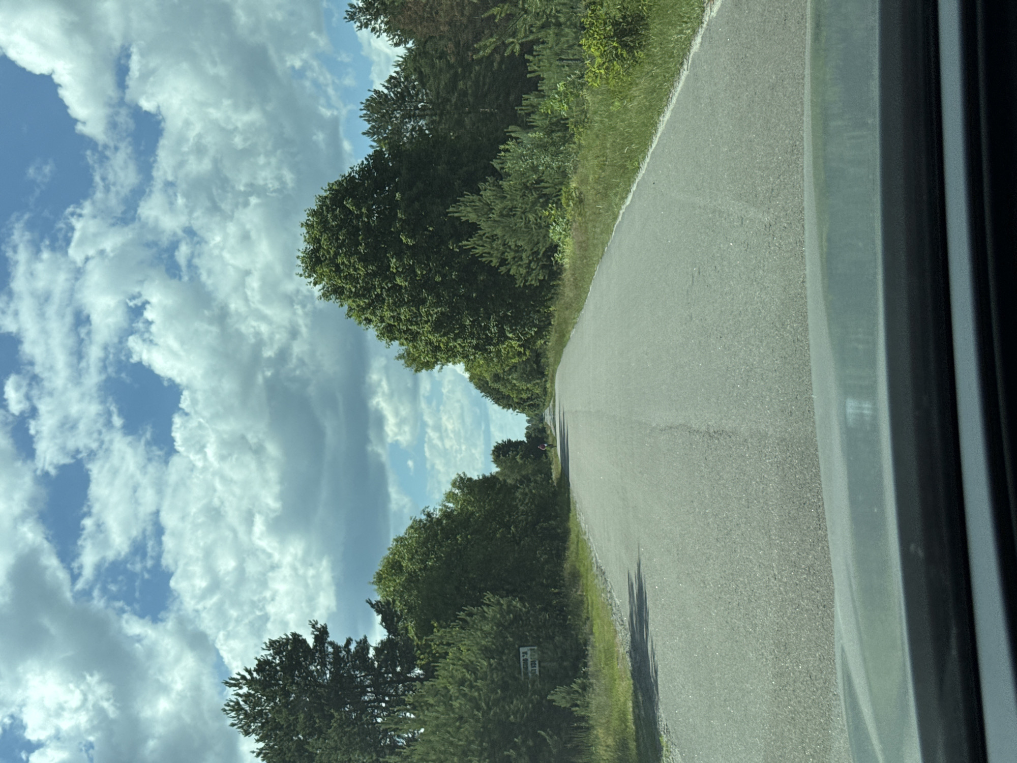 The image shows a paved road leading through a wooded area. The sky is partly cloudy with a mix of blue sky and white cumulus clouds. On the left side of the road, there is a sign. Further down the road, a person is visible on a bicycle. The surrounding vegetation consists of various trees and green grass. The perspective suggests the photo was taken from a vehicle.