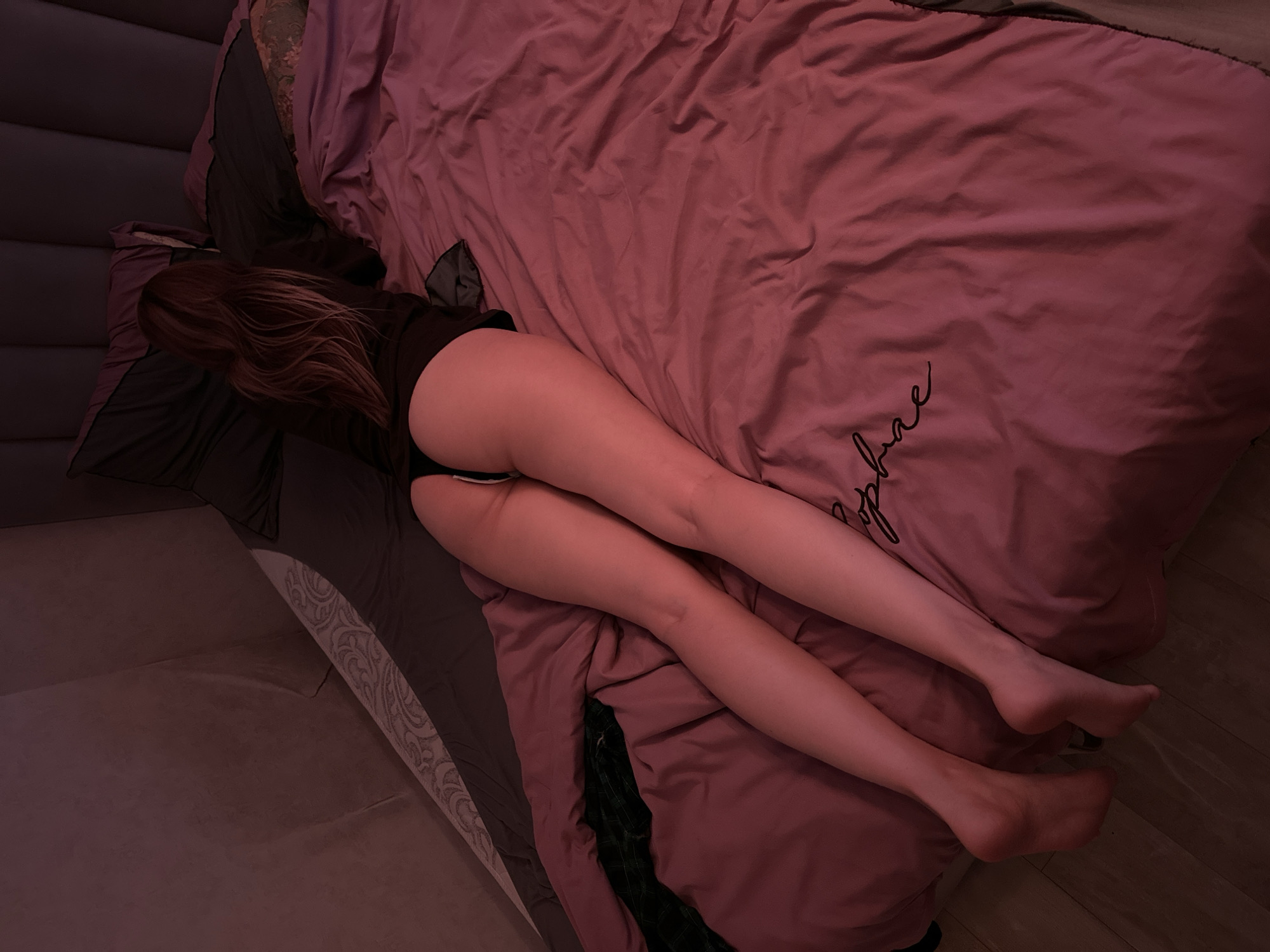 The image shows a person lying on a bed with their back to the camera. The person is wearing black underwear and a black top. The bed is covered with a pink comforter and pillows. The room is dimly lit, with pinkish light casting shadows. There is a headboard behind the bed. The person's legs are stretched out, and their feet are visible at the bottom of the frame. A script text is visible on the comforter. No specific details about the location in Naberezhnye Chelny, Russia, can be determined from the image.