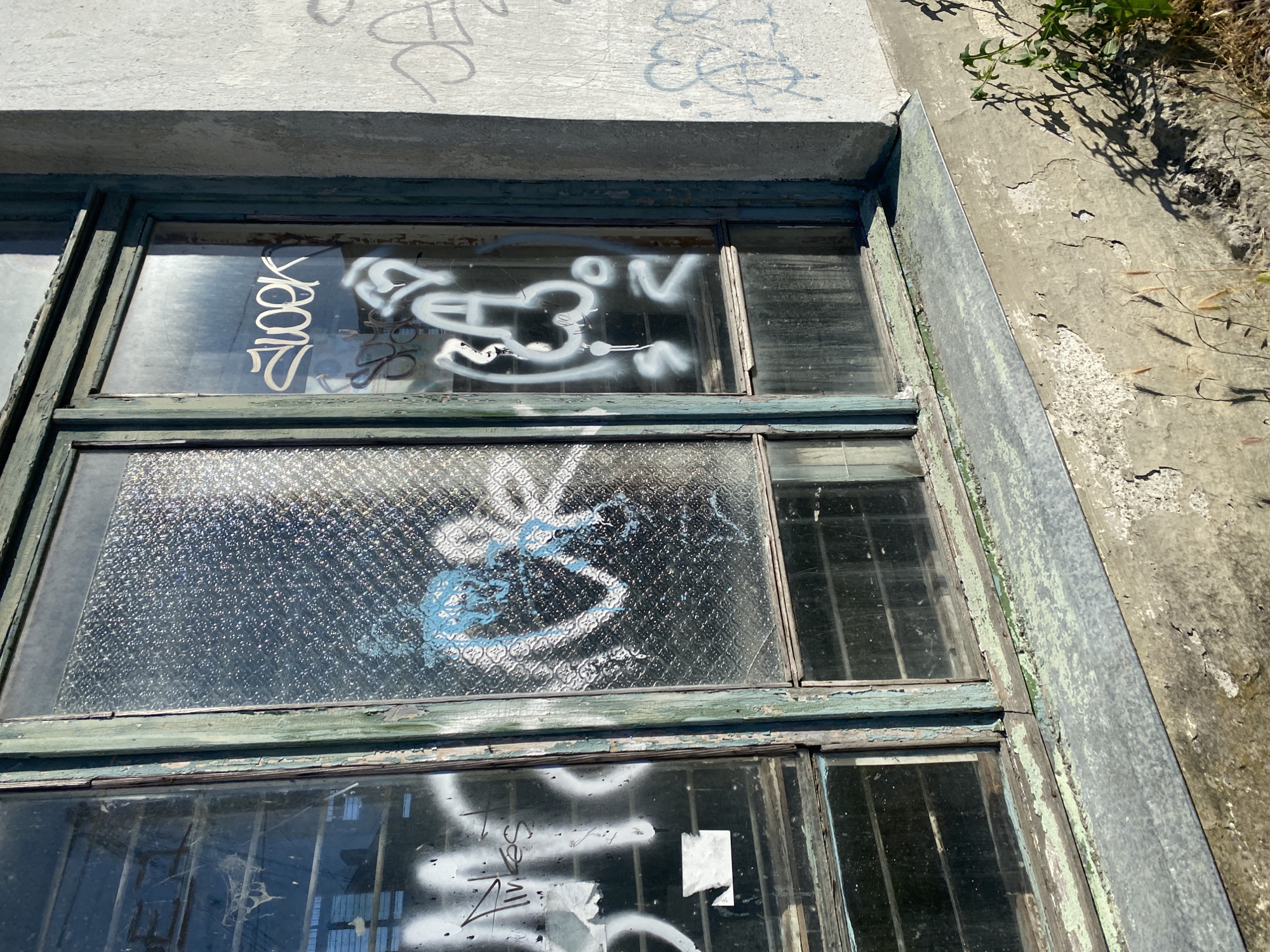 The image shows an exterior view of a weathered window frame covered in graffiti. The window is divided into multiple panes, some of which are clear and others are textured or barred. The window frame is painted a faded green, with visible peeling paint and rust. The surrounding wall is made of concrete and shows signs of age and decay, with cracks and flaking.
Graffiti in white and blue spray paint adorns the window panes and the concrete wall. Some of the graffiti is abstract, while other parts appear to be stylized lettering or symbols. Visible text includes "2LOKK" written vertically on the left pane of the top window, and a circular symbol with "3" and "N" on the right pane. Further down, another pane has what appears to be a stylized "S" in blue. The bottom pane also has white spray paint, with some illegible text and what looks like a ripped piece of paper attached.
The scene is outdoors, under bright sunlight, suggesting a daytime setting. The presence of dry grass and weeds growing in the upper right corner indicates an unmaintained outdoor area. The overall impression is one of urban decay and neglect, with the graffiti adding a layer of artistic expression or vandalism to the weathered surfaces. The location context of Sevastopol, Ukraine, suggests a possibly post-Soviet era building or a location experiencing urban blight. No people are visible in the image.