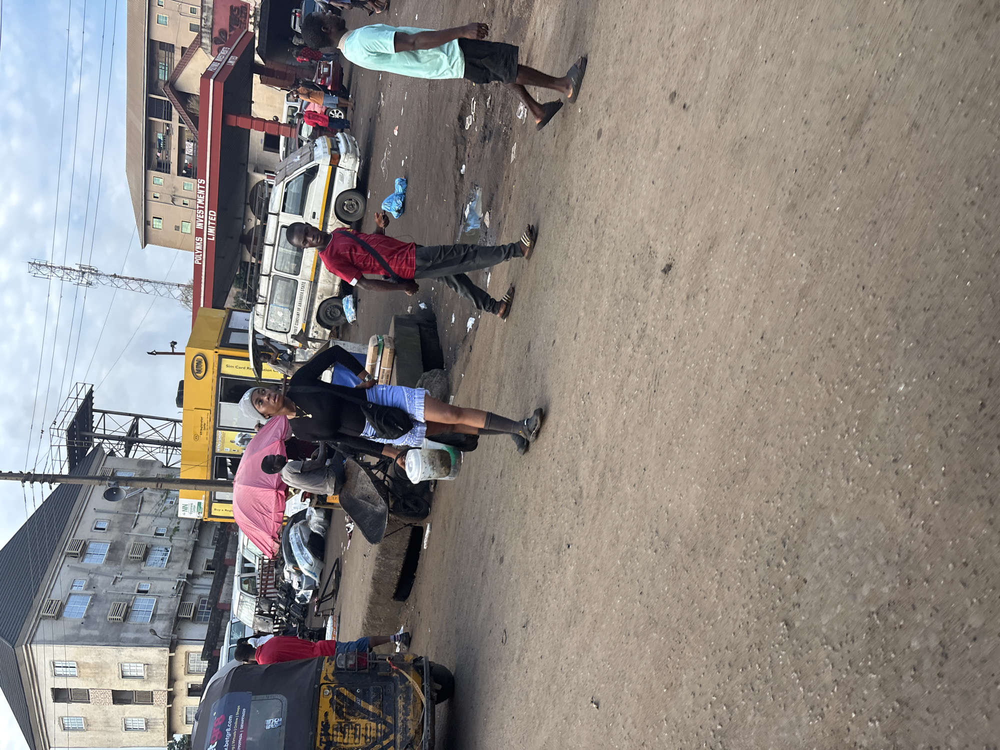 The image depicts a street scene in Onitsha, Nigeria, likely a market area given the presence of various vehicles and people engaged in activities. A prominent feature is a row of buildings, one of which appears to be a gas station with the sign "POLYNKS INVESTMENTS LIMITED." Several vehicles, including a three-wheeled auto-rickshaw and vans, are present. A pink umbrella is open, suggesting it is a stall or shade.
In the foreground, a woman wearing a black top, blue skirt, and gray socks is walking and carrying two plastic containers. To her left, a man in a red shirt and dark pants is walking away from the camera. To the right, another man in a light green t-shirt and black shorts is walking towards the right side of the frame. The ground is paved with a dusty, uneven surface, with scattered debris and puddles of water visible. The overall environment appears to be an active commercial or transportation hub within the city.