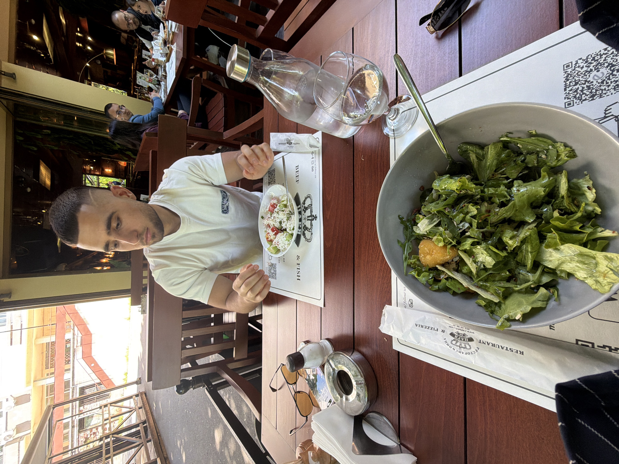 The image depicts a man sitting at an outdoor table in a restaurant, likely located in Stara Zagora, Bulgaria. The table is wooden and holds a large bowl of salad, a small bowl of another salad, a water bottle with a glass, and an ashtray with sunglasses and napkins. There is also a printed placemat with the words "RESTAURANT" and "PIZZERIA". The man is wearing a white t-shirt. Other people are faintly visible seated at a table in the background. The outside environment shows buildings across the street.