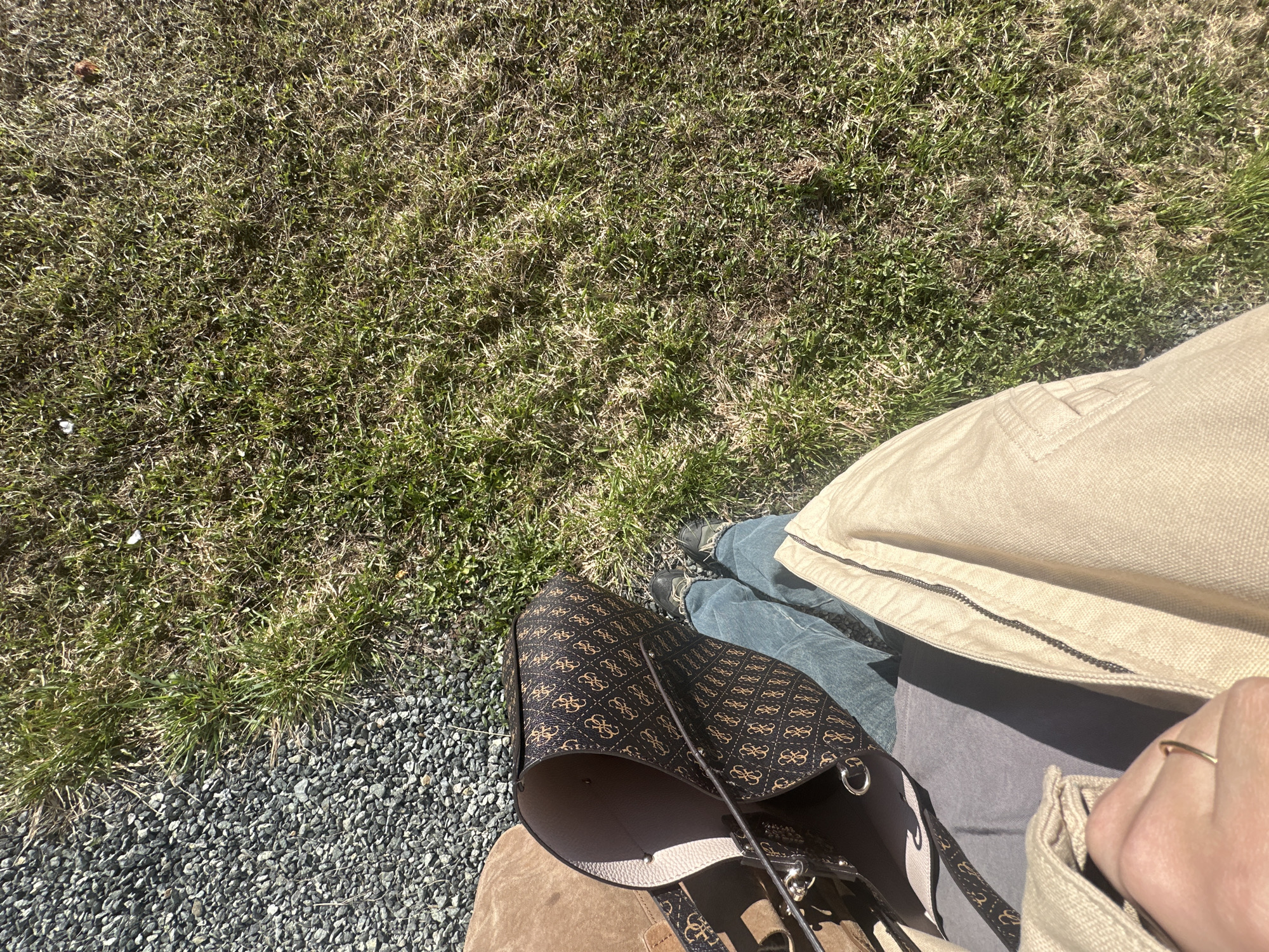 A high-angle outdoor shot depicts a person standing on gravel next to a grassy area. The person is wearing denim jeans, a beige jacket, and dark-colored sneakers. A brown and black monogrammed tote bag sits adjacent to the person on the gravel. The location cannot be determined to be Kharnavy, Russia from the image.