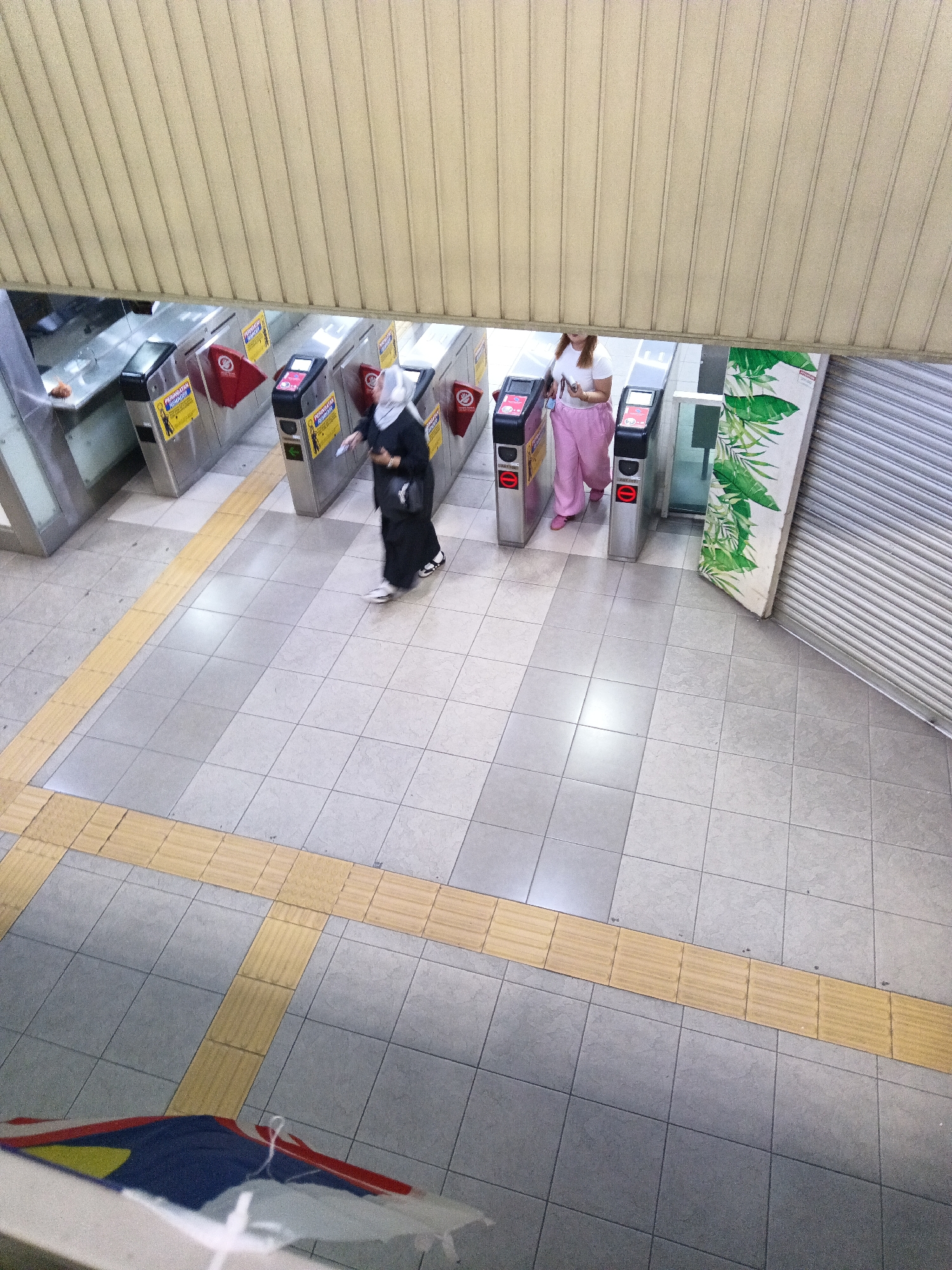 The image captures a high-angle view of an indoor environment, likely a public transportation station, with two women interacting with ticket gates. One woman, dressed in a black abaya and hijab, is seen swiping a card or ticket at a gate. The other woman, wearing a white t-shirt and pink pants, is standing near a gate, holding a mobile phone. The gates are metallic and have yellow signage with red warning symbols. The floor is tiled in a grid pattern, with yellow tactile paving strips indicating a pathway. Above the gates, a beige, ribbed ceiling extends across the frame, and to the right, a rolled-up metal shutter and a wall with a tropical plant motif are visible. The scene suggests a typical urban transit point in Kuala Lumpur, Malaysia, characterized by its modern infrastructure and bustling pedestrian activity.