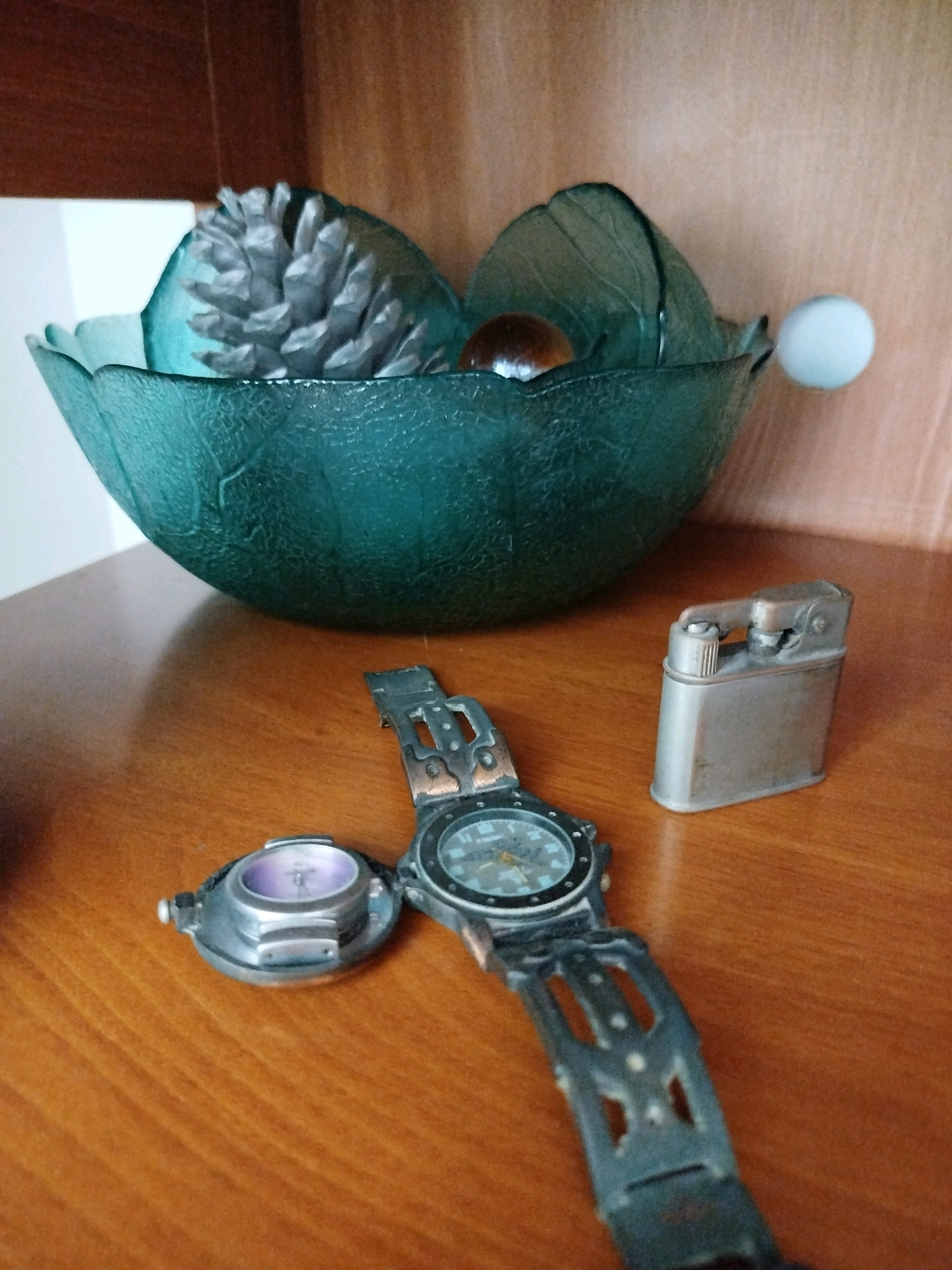 The image shows a close-up, eye-level view of a wooden surface with several objects placed upon it, likely indoors. In the background, a shelf with a dark wood finish is visible, holding a translucent teal-colored bowl filled with decorative items. The bowl contains what appears to be a silver-painted pinecone, a small brown sphere, and two green, leaf-shaped decorative pieces. A small white sphere is attached to the right side of the bowl.
In the foreground, on the wooden surface, are three distinct objects. To the left, a small, silver-colored compass or navigational device with a purple-tinted display is open. To the right of the compass, a man's wristwatch with a black band and a dark dial is laid out. The band appears to be made of a textured material with cut-out sections. To the right of the wristwatch, a silver-colored metal lighter is positioned upright. The overall scene suggests a still life composition, possibly a collection of personal items. There is no indication of the location being in Ashkelon, Israel.