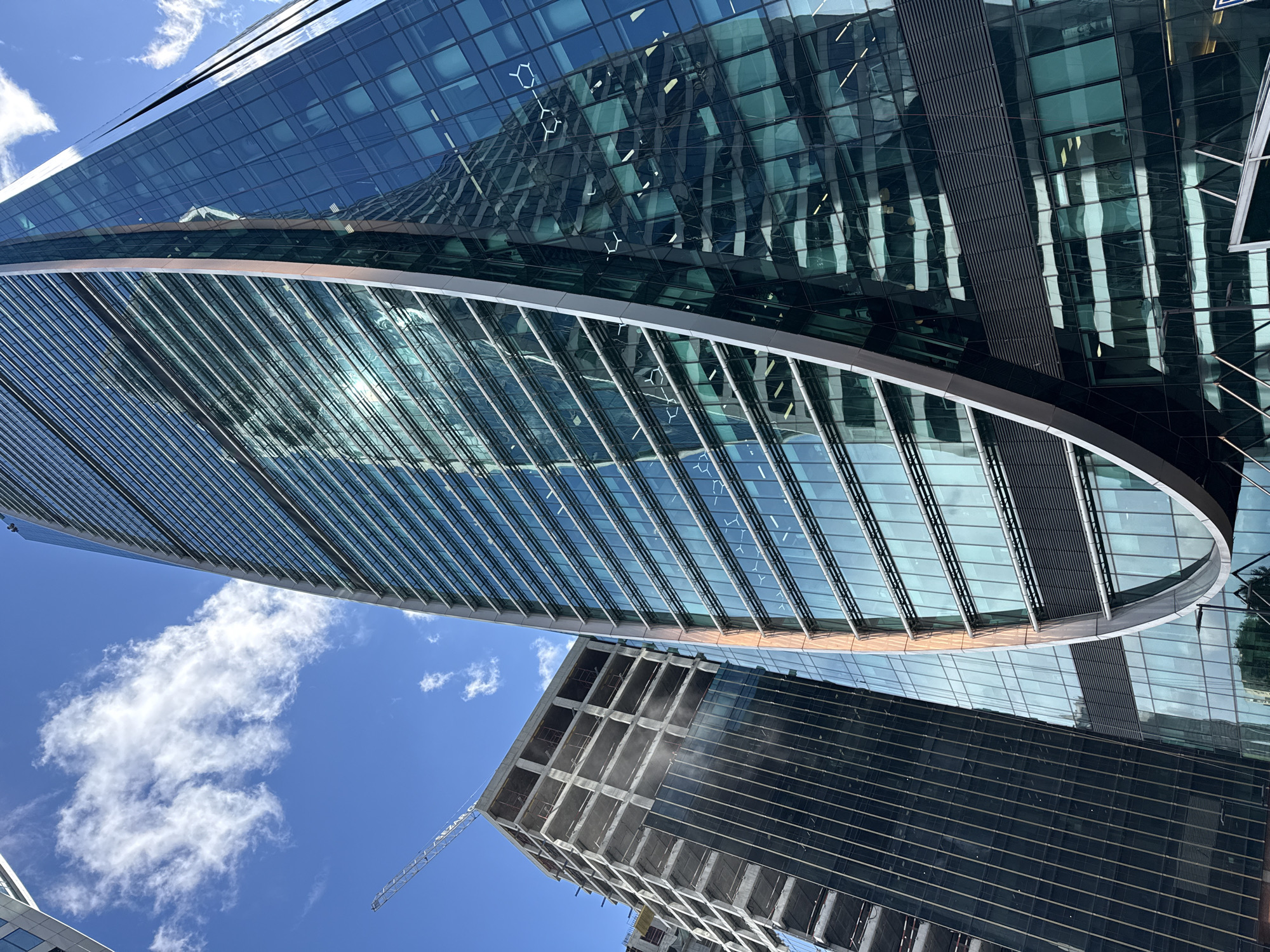 The image shows a low-angle view of modern skyscrapers in Moscow, Russia. On the right, a sleek, curved skyscraper covered in reflective blue glass dominates the frame. Its facade features horizontal bands of windows and metallic accents, with reflections of other buildings and the sky visible. To the left, a building under construction is partially visible, with its concrete framework exposed and dark glass panels on its lower section. A construction crane extends into the bright blue sky, which is dotted with scattered white clouds. The overall scene depicts urban development and architectural modernity.