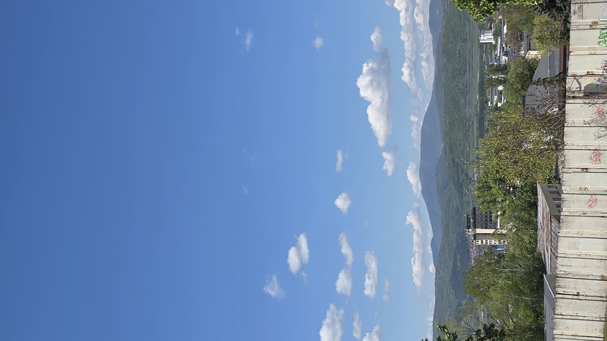 The image shows a view in Blagoevgrad, Bulgaria. A large mountain is present. The sky is blue with scattered clouds. A cityscape is visible, obscured by trees, along with buildings and metal containers.
